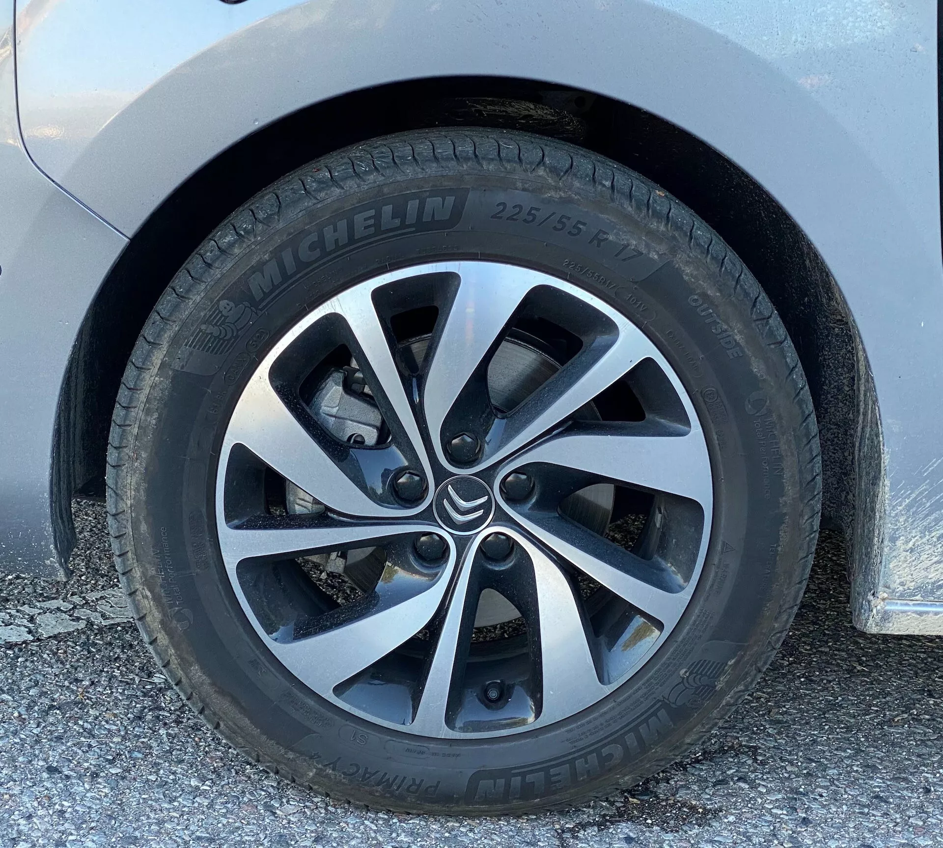 Zbliżenie koła Citroena Spacetourera 2016 z niskiego kąta, niemal na poziomie gruntu, podkreślające oponę marki Michelin (225/55) i nowoczesne felgi aluminiowe z pięcioma podwójnymi szprychami. Koło jest czyste, z minimalnymi zabrudzeniami, ukazując część systemu hamulców tarczowych przez szprychy. Nad kołem znajduje się karoseria jasnego koloru, prawdopodobnie biała lub srebrna.