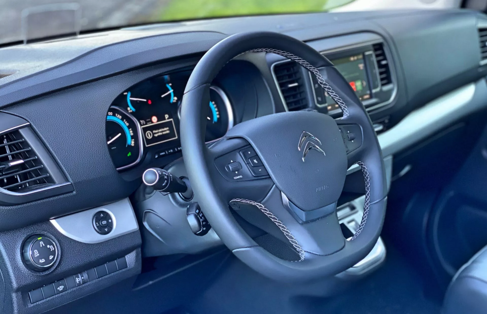 Widok wnętrza Citroen Spacetourer 2016 skupiający się na obszarze kierowcy, pokazujący kierownicę z zamontowanymi kontrolkami, zestaw wskaźników z analogowymi zegarami i cyfrowym wyświetlaczem oraz częściowo widoczny ekran centralny i przyciski klimatyzacji na środkowej konsoli. Widoczne są również elementy sterujące szybami i lusterkiem bocznym z funkcjami elektrycznej regulacji na drzwiach kierowcy.