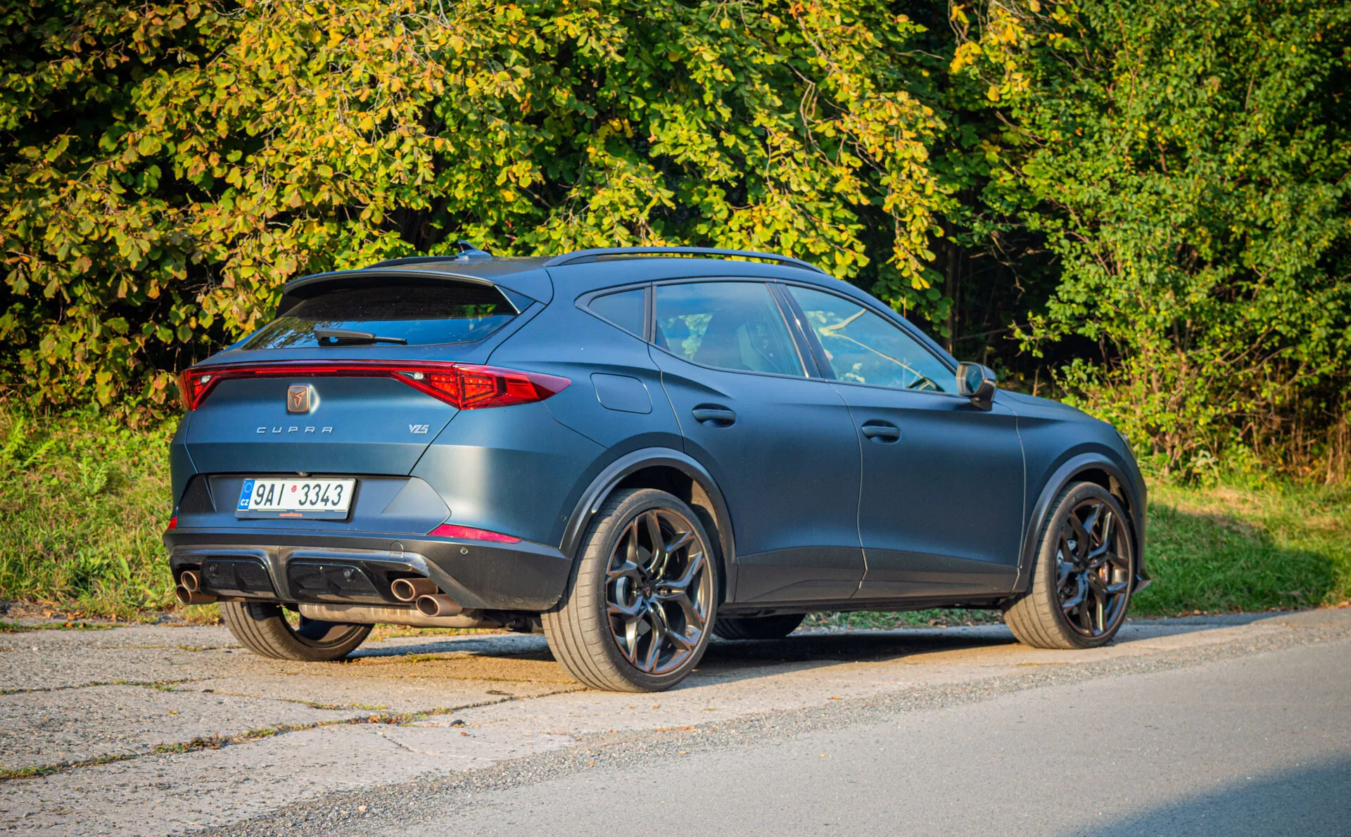 Nowoczesny ciemnoszary matowy czarny crossover SUV Cupra Formentor 2020 zaparkowany na asfalcie, prezentujący widok z tyłu trzy czwarte podkreślający design typu coupé, sportową aerodynamikę, tylne światła LED, tylny spoiler, podwójne rury wydechowe i duże koła ze stopów w scenerii przedmieścia