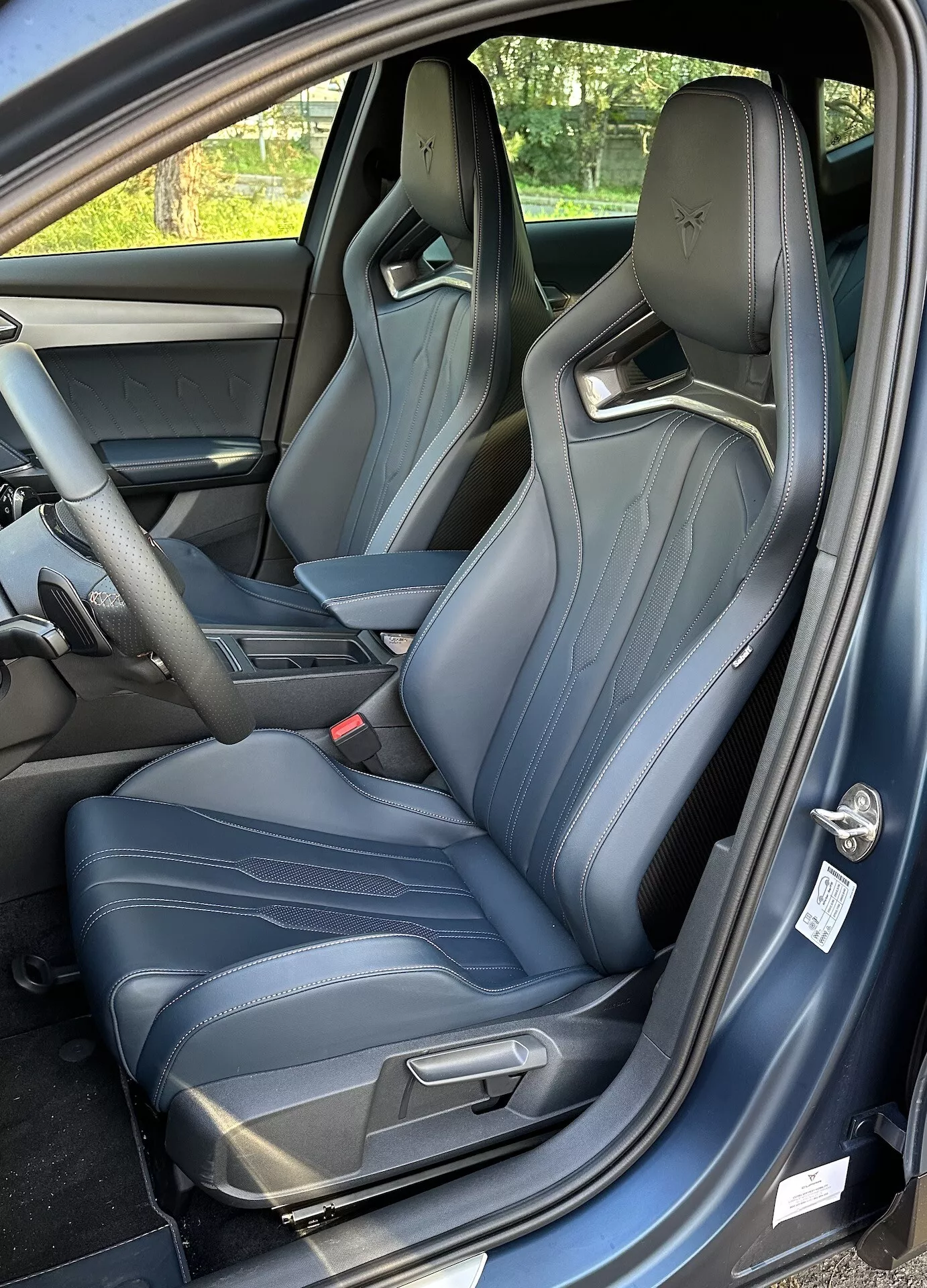 Widok wnętrza Cupra Formentor 2020, pokazujący fotel kierowcy z perspektywy trzy czwarte, z otwartymi drzwiami, sportowymi siedzeniami kubełkowymi z jasnoniebieskim szwem i wglądem na konsolę centralną i deske rozdzielczą.