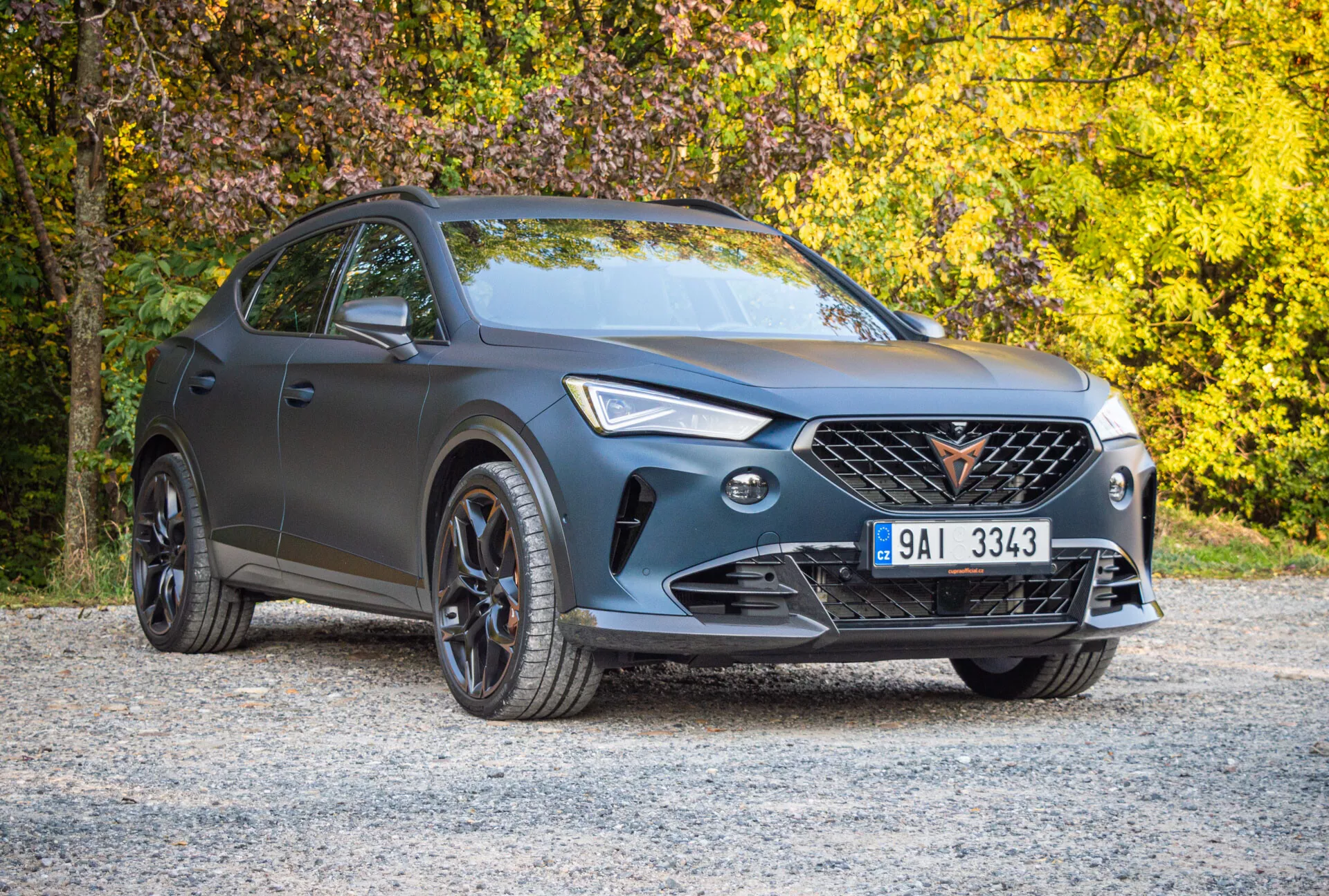 Cupra Formentor z 2020 roku zaparkowana na żwirowej nawierzchni z jesiennym liściem w tle, prezentująca matowe szare wykończenie, charakterystyczną atrapę chłodnicy, reflektory LED i sportowe czarne wykończenia