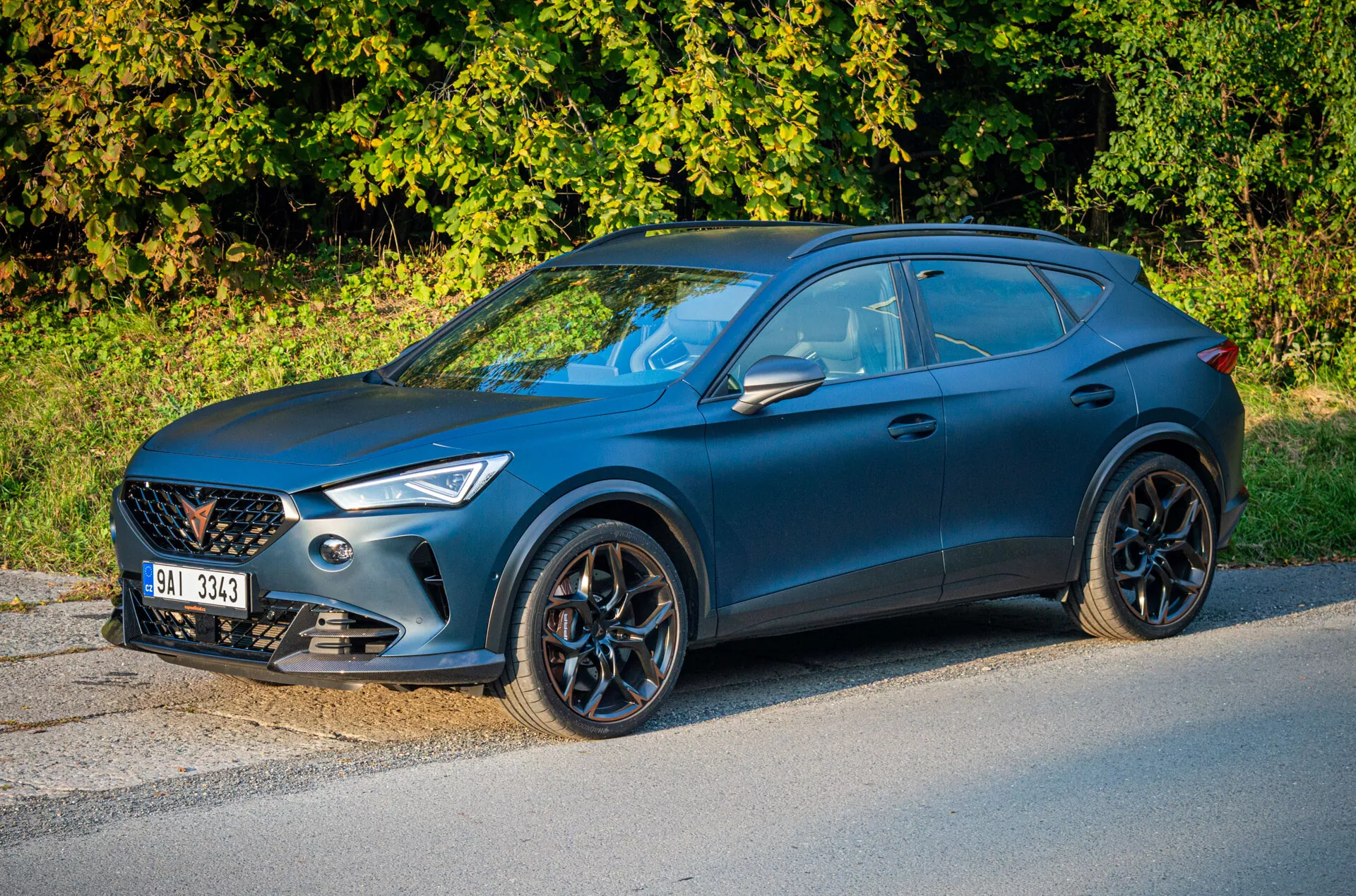 Niebieskie crossover SUV Cupra Formentor 2020 zaparkowane przy drodze z roślinnością w tle, podkreślające widok z przodu z trzech czwartych skupiony na stronie kierowcy, grill przedni, kątowe reflektory, wieloramienne alufelgi i sportowy profil boczny typu coupe