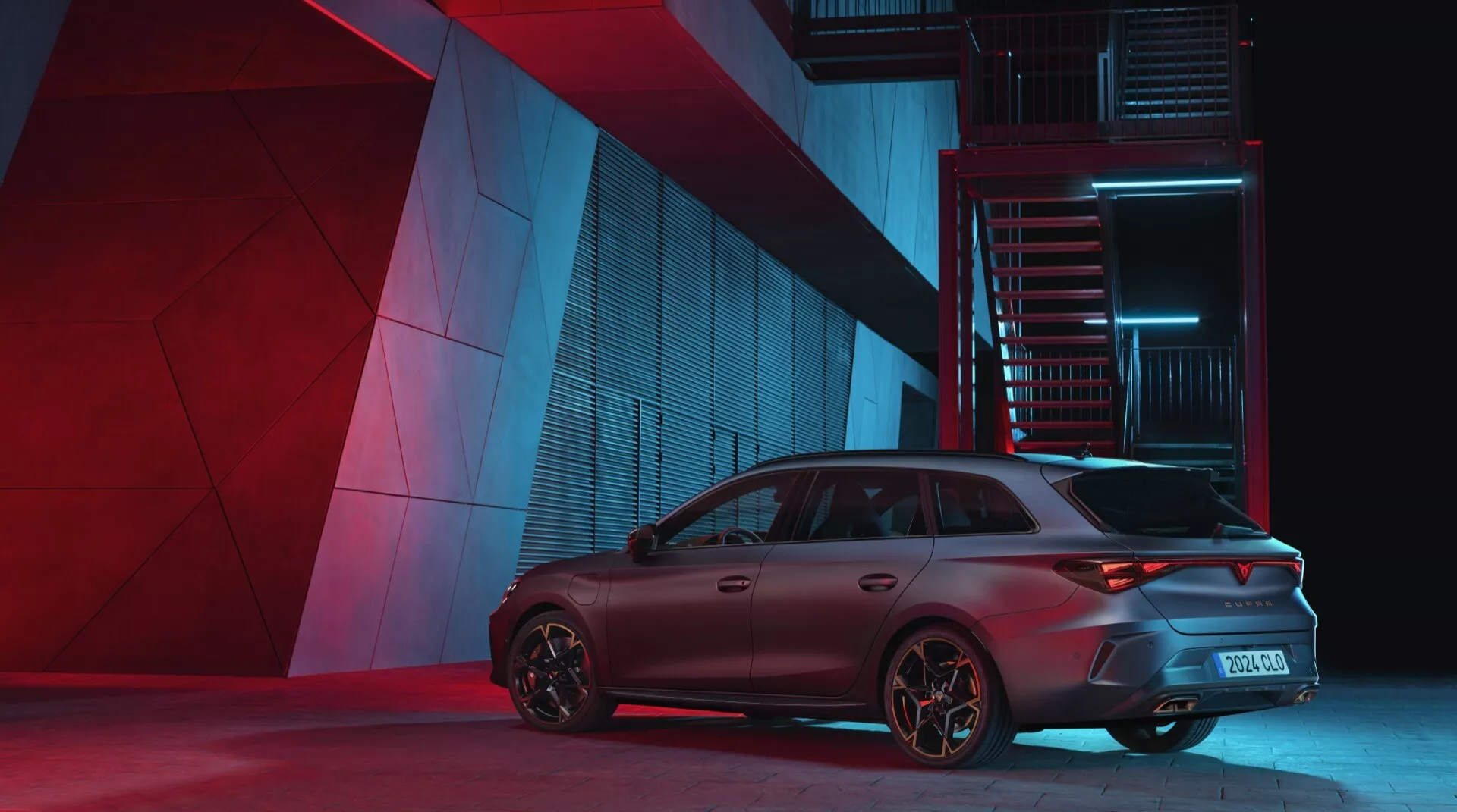 Cupra Leon 2024 pierwsza generacja po liftingu, sfotografowana pod kątem tylno-trzyczwardziowym, podkreślającym stronę pasażera. Dramatyczne nocne oświetlenie podkreśla cechy projektowe samochodu, w tym sportowe światła tylne, tylny zderzak, agresywny spojler dachowy i felgi zorientowane na osiągi na tle miejskiego przemysłowego otoczenia.