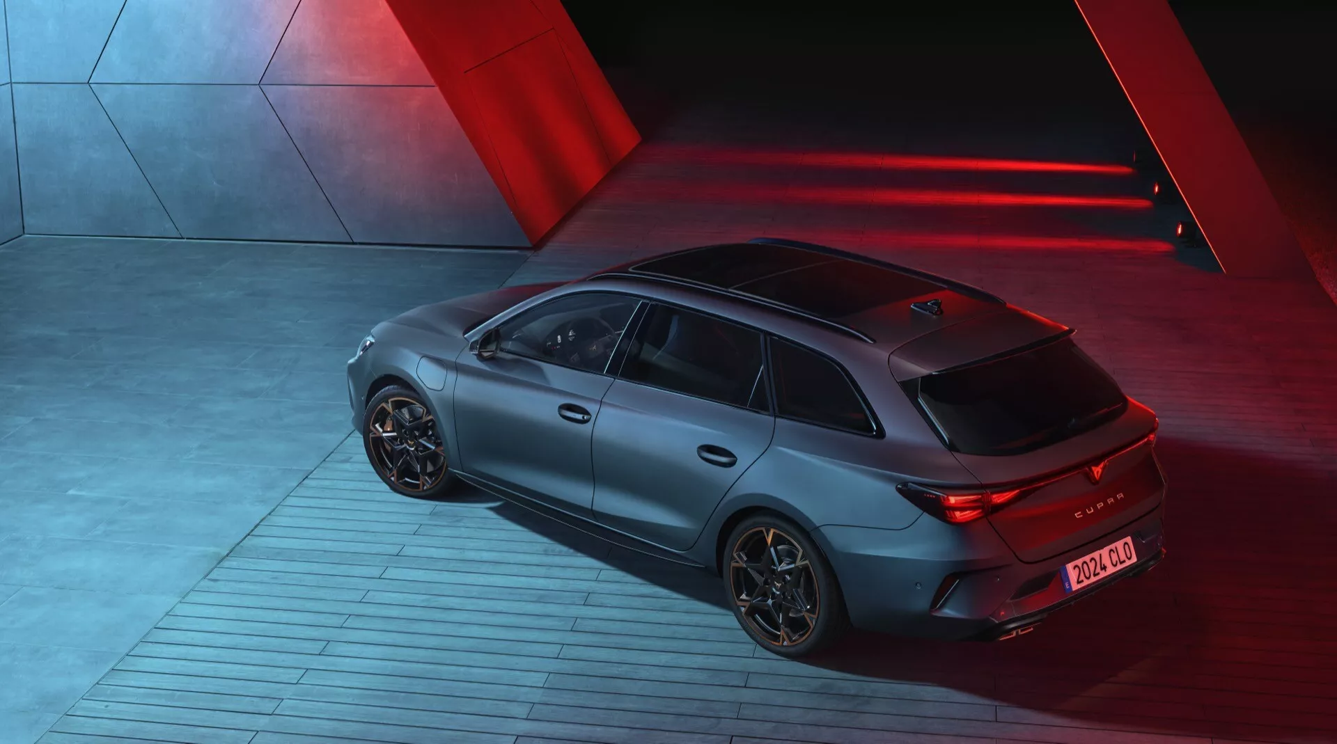 Widok z góry na Cupra Leon 2024 (1. generacja lifting) zrobiony w nocy, prezentujący tył oraz prawy bok sportowego hatchbacka. Samochód jest oświetlony dramatycznymi czerwonymi światłami kierunkowymi, tworzącymi elegancki, futurystyczny klimat. Charakteryzuje się matowym wykończeniem, podświetlanymi tylnymi światłami LED i aerodynamicznymi felgami aluminiowymi. Pojazd stoi na wzorzystej industrialnej powierzchni, sugerując ruch. Widoczne elementy technologiczne obejmują tylny dyfuzor oraz krótką antenę z tyłu dla technologii komunikacyjnych.