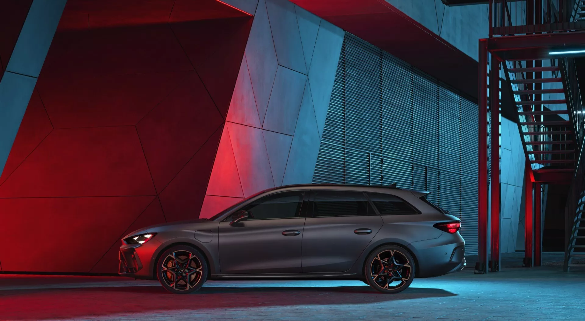 Widok z profilu bocznego sportowego i eleganckiego samochodu Cupra Leon 2024 w miejskim środowisku nocnym z sztucznym czerwonym i niebieskim oświetleniem w tle. Samochód charakteryzuje się długą, aerodynamiczną sylwetką z przyciemnianymi szybami i chromowanymi akcentami, linią dachu kończącą się spojlerem i charakterystycznymi wieloszprychowymi felgami aluminiowymi. Widoczne zaciski hamulcowe sugerują hamulce nastawione na osiągi. Karoseria ma konturowane linie, a z tego kąta żadne znaki ani cechy technologiczne nie są łatwo identyfikowalne.