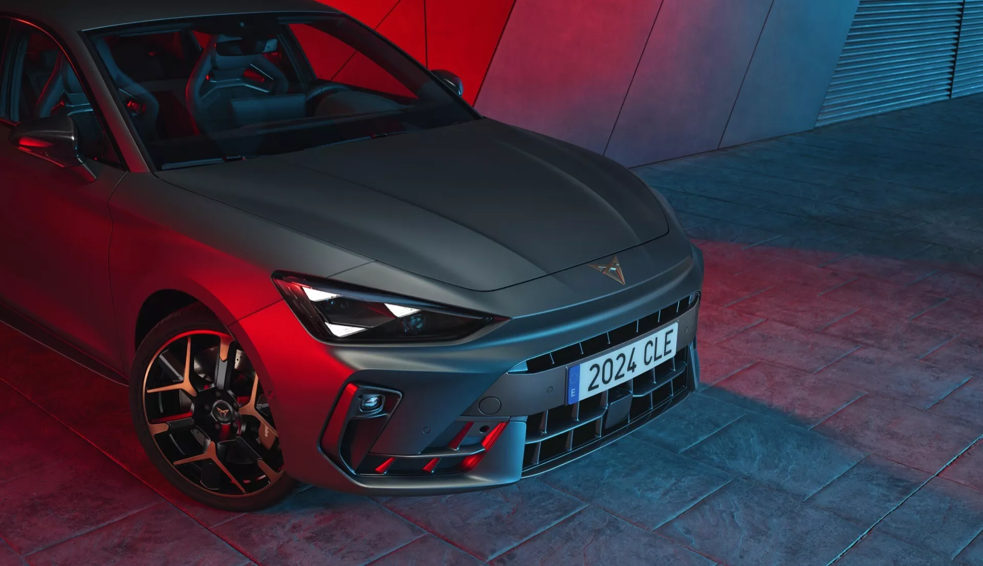 Widok z niskiego przodu na Cupra Leon 2024 (pierwsza generacja facelift), prezentujący cechy projektowe z wyraźnym grillem, eleganckimi reflektorami z LED-owymi światłami do jazdy dziennej, sportowym zderzakiem z wlotami powietrza oraz wydajnymi felgami aluminiowymi z wysokowydajnymi zaciskami hamulcowymi. Metaliczna barwa lakieru i dramatyczne czerwone oświetlenie otoczenia sugerują wysokiej klasy pojazd nastawiony na wydajność, prawdopodobnie sfotografowany na podłodze salonu.