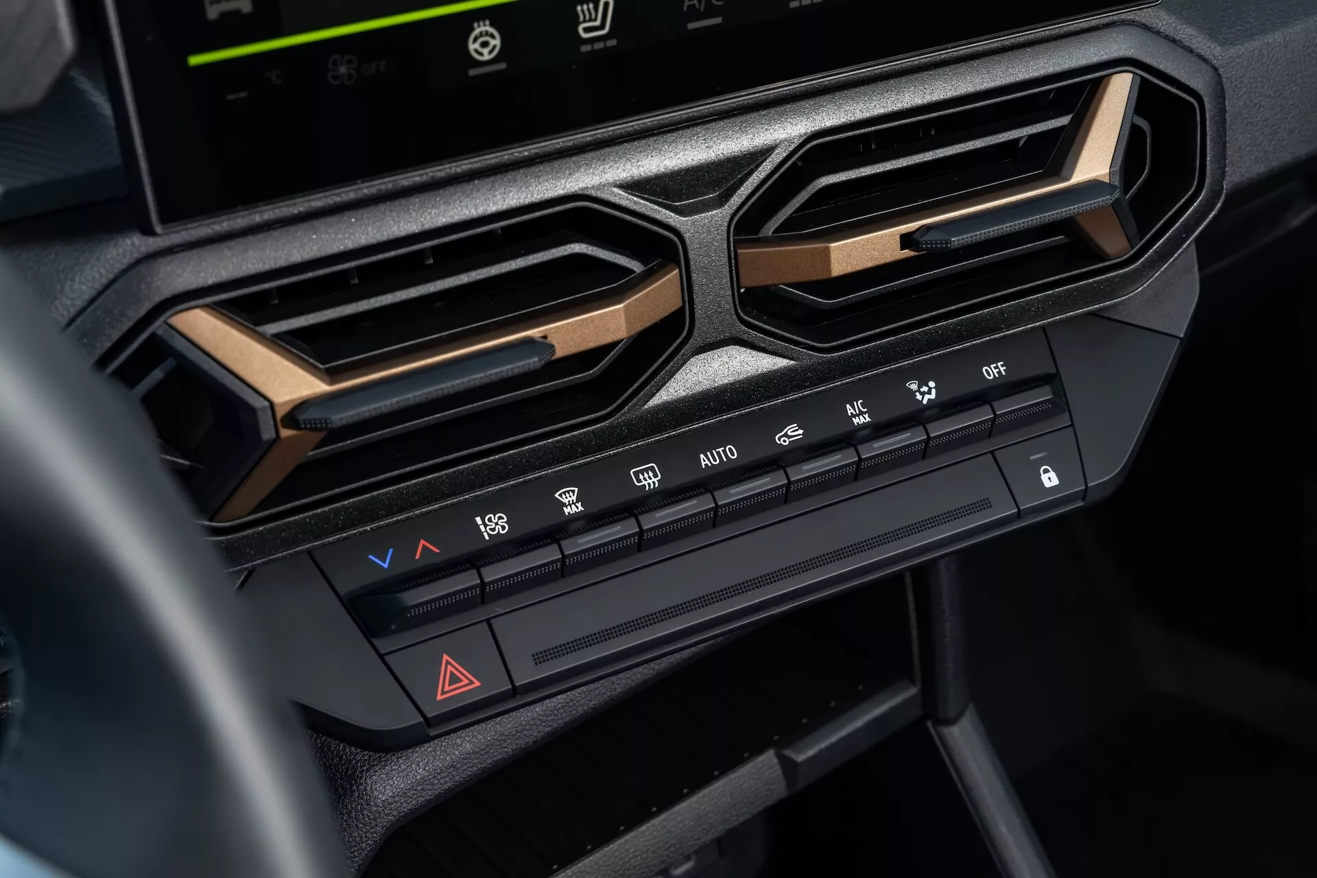 Zbliżenie systemu kontroli klimatu w Dacia Duster 3. generacji 2024 na centralnym desce rozdzielczej pod ekranem systemu rozrywki, podkreślające regulacje temperatury i prędkości wentylatora, funkcje dystrybucji powietrza, funkcje odmrażania, przycisk świateł awaryjnych oraz opcje klimatyzacji, oprawione przez kątowe nawiewy i akcentowane metalicznym wykończeniem dla nowoczesnej estetyki wnętrza
