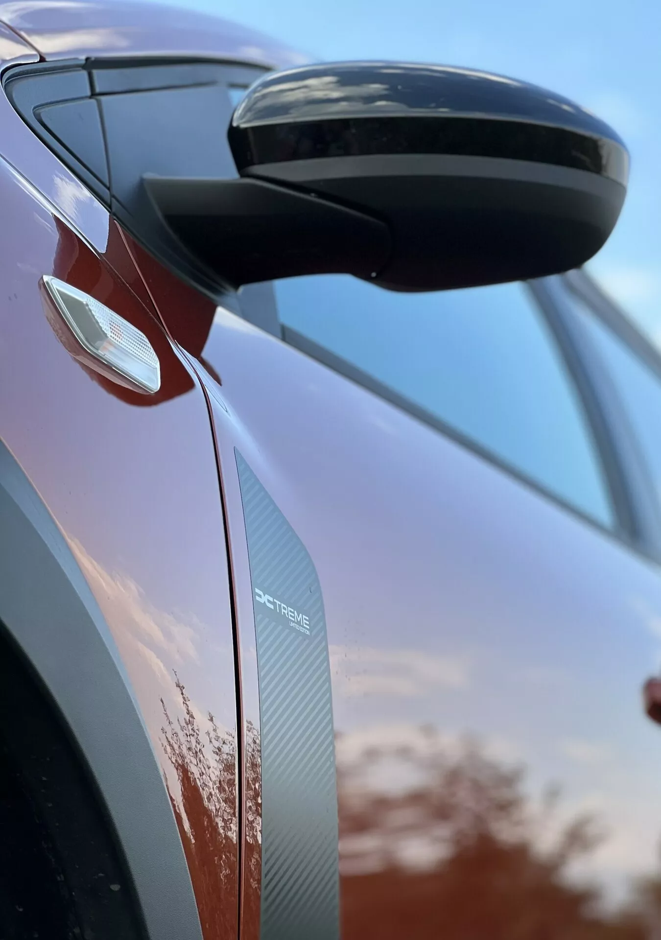 Zbliżony detal Dacia Jogger 2022 skupiający się na bocznym lusterku wstecznym i projekcie drzwi, prezentujący cechy aerodynamiczne, zintegrowane kierunkowskazy i tekstury z włókna węglowego na tle nieba