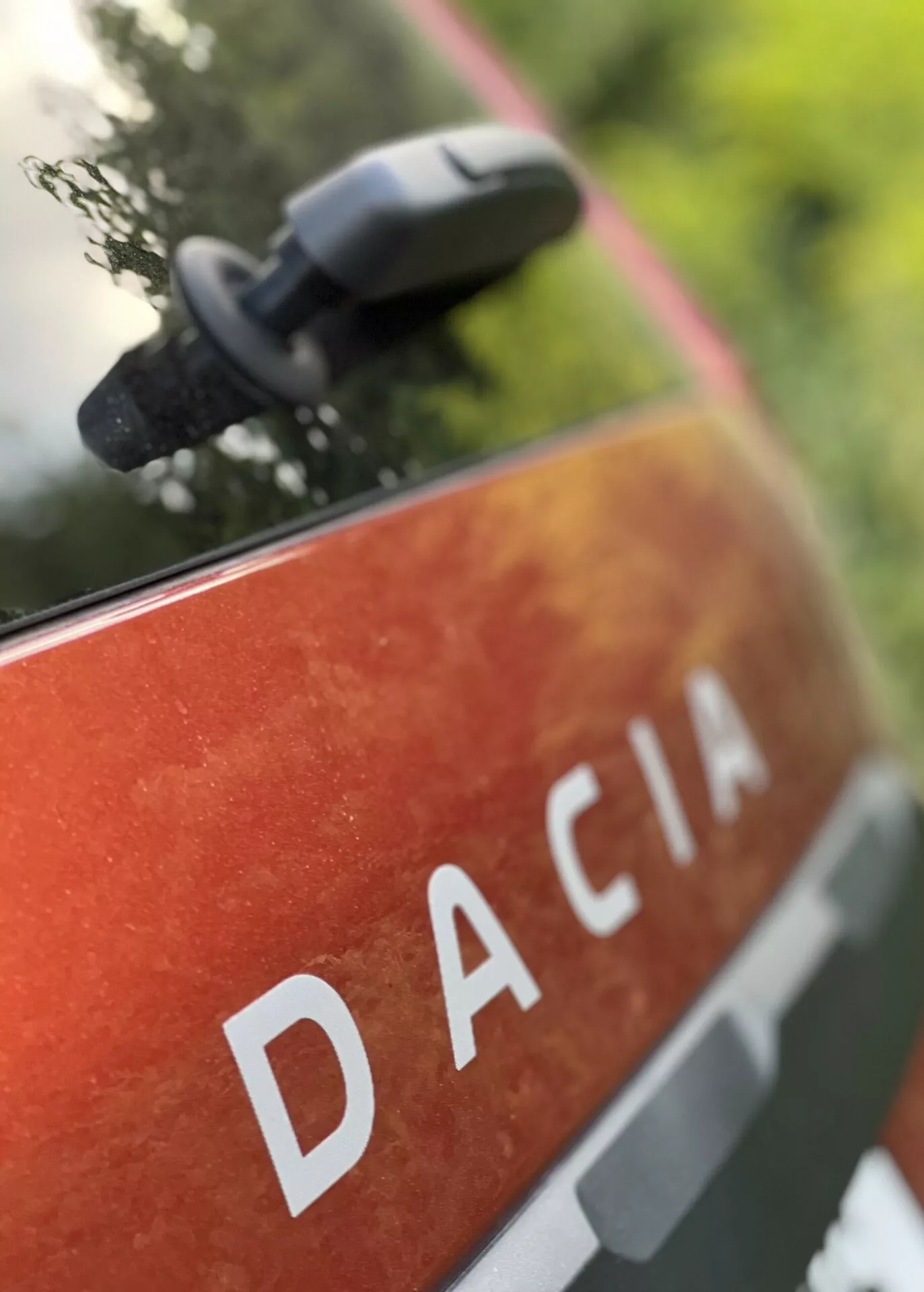 Zbliżenie na tylny panel Dacia Jogger 2022 z napisem DACIA dużymi literami na metaliczno-pomarańczowo-czerwonym lakierze z plamami i brudem, sugerującym użytkowanie zewnętrzne i trwałość, umieszczone w naturalnym środowisku z rozmazanym lusterkiem bocznym wskazującym na nowoczesne funkcje lustra.