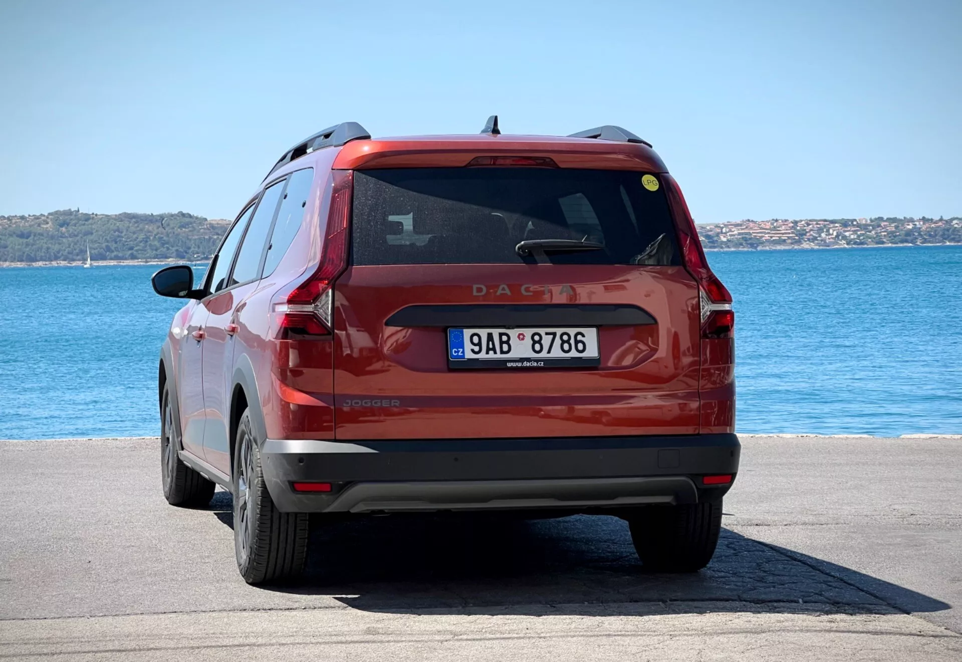 Tylny widok czerwonego kompaktowego SUV-a Dacia Jogger 2022 zaparkowanego na wybrukowanym obszarze przybrzeżnym pod jasnym światłem dzienym, prezentujący cały tył pojazdu, w tym tylne światła, tylną szybę i tablicę rejestracyjną na tle niebieskiej wody i czystego nieba.