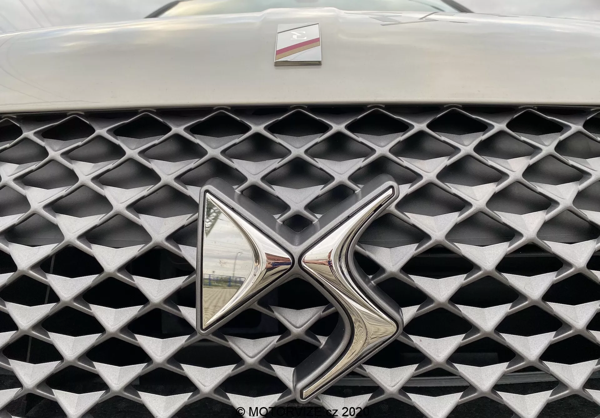 Zbliżenie na grille DS Automobiles DS 3 Crossback E-Tense 2019. Obiekt uwiecznia grille z perspektywy frontalnej, skupiając się na wysoce odblaskowym metalowym emblemacie. Grille ma charakterystyczną teksturę o diamentowym wzorze w matowym szarym wykończeniu, kontrastującym z błyszczącym chromowanym emblematem. Ograniczone pole widzenia poza grillem i emblematem, tło rozmyte.