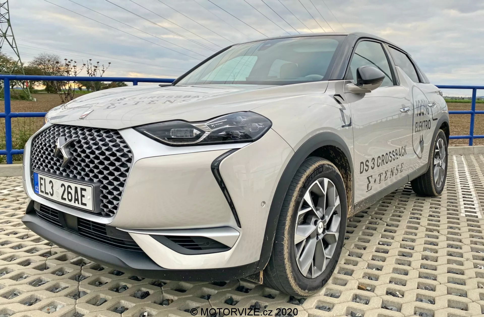 Na zdjęciu widoczny jest nowoczesny SUV crossover DS-Automobiles DS-3 2019 (DS-3 Crossback E-Tense), uchwycony pod kątem przednim na powierzchni złożonej z przeplatających się płytek. Perspektywa z niskiej do średniej wysokości podkreśla sylwetkę samochodu, zapewniając kompleksowy widok na jego przednią kratę z charakterystycznym geometrycznym wzorem, reflektory LED, felgi aluminiowe, lusterka boczne oraz detale marki. Długość i cechy konstrukcyjne przodu pojazdu są wyraźnie widoczne, podkreślając nowoczesną i zaawansowaną technologię motoryzacyjną.