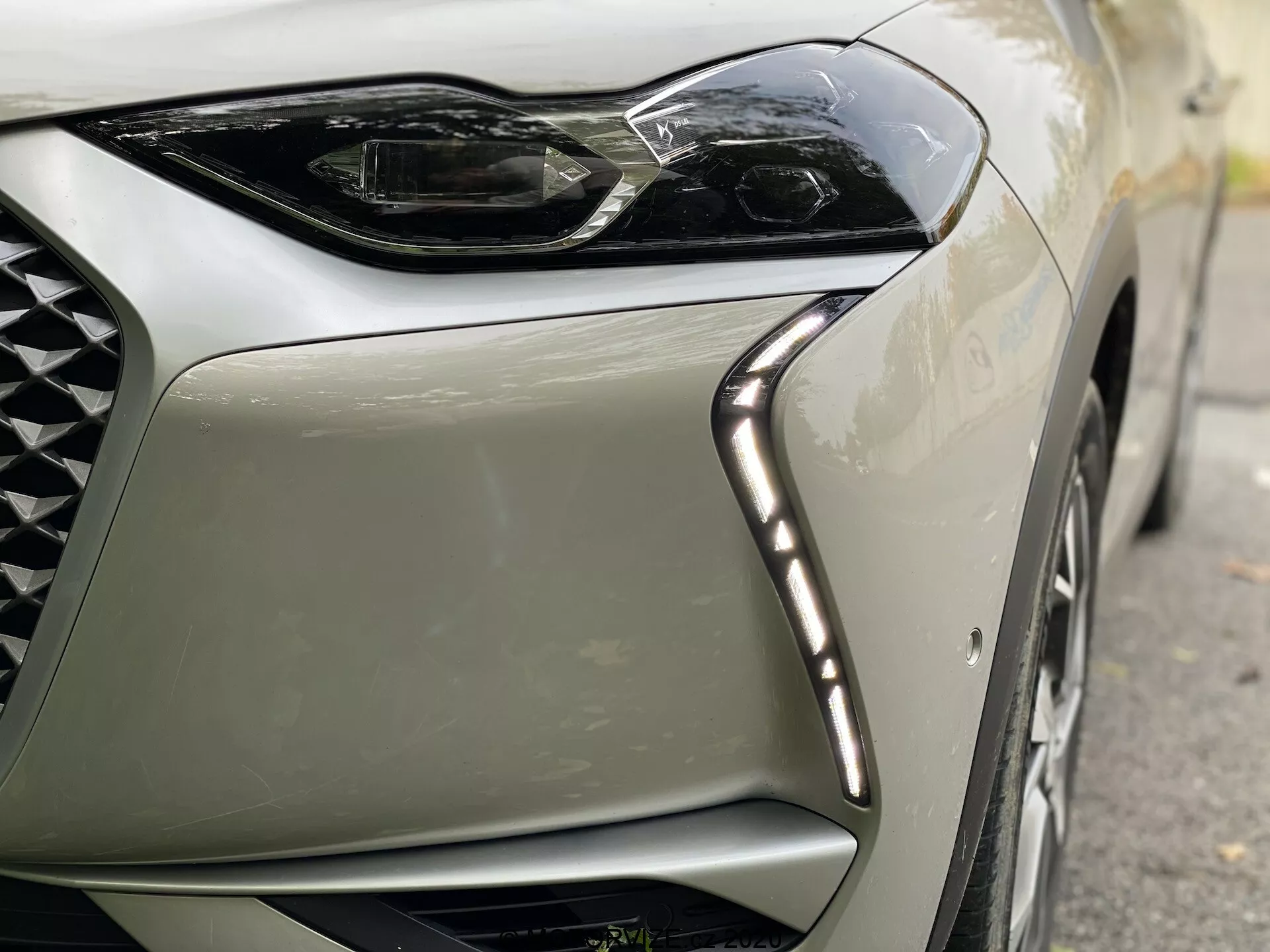 Zbliżenie przedniego lewego rogu nowoczesnego samochodu DS Automobiles DS-3 Crossback E-Tense z 2019 roku. Obraz koncentruje się na eleganckim, kanciastym wzornictwie reflektorów LED z dziennymi światłami LED wzdłuż górnej krawędzi oraz części kratki o wzorze diamentowym, podkreślając luksus i nowoczesną technologię.