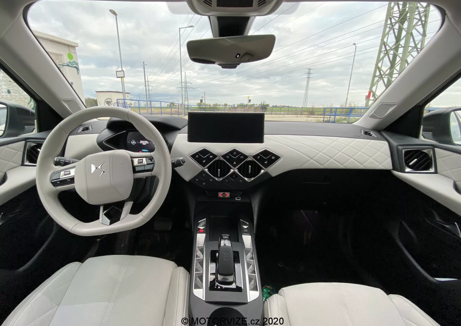 Widok z tylnego siedzenia na nowoczesne wnętrze centralne DS Automobiles DS-3 Crossback E-Tense 2019. Zdjęcie uchwyca trójramienną kierownicę z zamontowanymi przyciskami, cyfrowy zestaw wskaźników, dużą dotykową konsolę na środku z sześciokątnymi przyciskami sterowania klimatem poniżej oraz dźwignię zmiany biegów sugerującą automatyczną skrzynię biegów. Deska rozdzielcza i drzwi mają wzór pikowania, wykonany z ekoskóry lub skóry, a przednie fotele kubełkowe są tapicerowane jasną skórą lub syntetycznym materiałem. Widoczne cechy to podświetlenie ambientowe wzdłuż krawędzi, panoramiczny dach i widoczne otwory wentylacyjne na desce rozdzielczej.