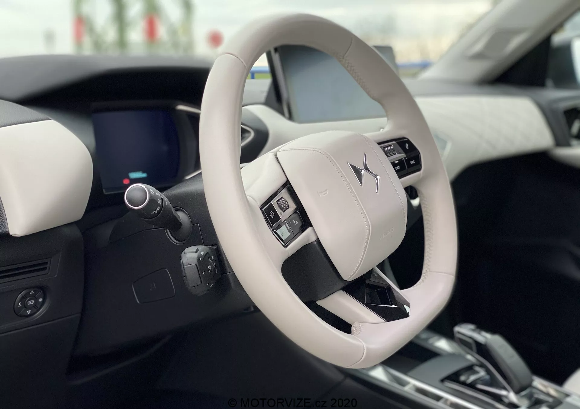Widok wnętrza DS Automobiles DS 3 Crossback E-Tense 2019, ukazujący nowoczesny i luksusowy design kierownicy i deski rozdzielczej z perspektywy fotela kierowcy. Jasnokolorowa kierownica z przyciskami sterowania wyraźnie eksponuje logo producenta. Elegancki cyfrowy zestaw wskaźników, częściowo widoczny za kierownicą, uzupełnia dwukolorową stylistykę wnętrza z wysokiej jakości materiałami.