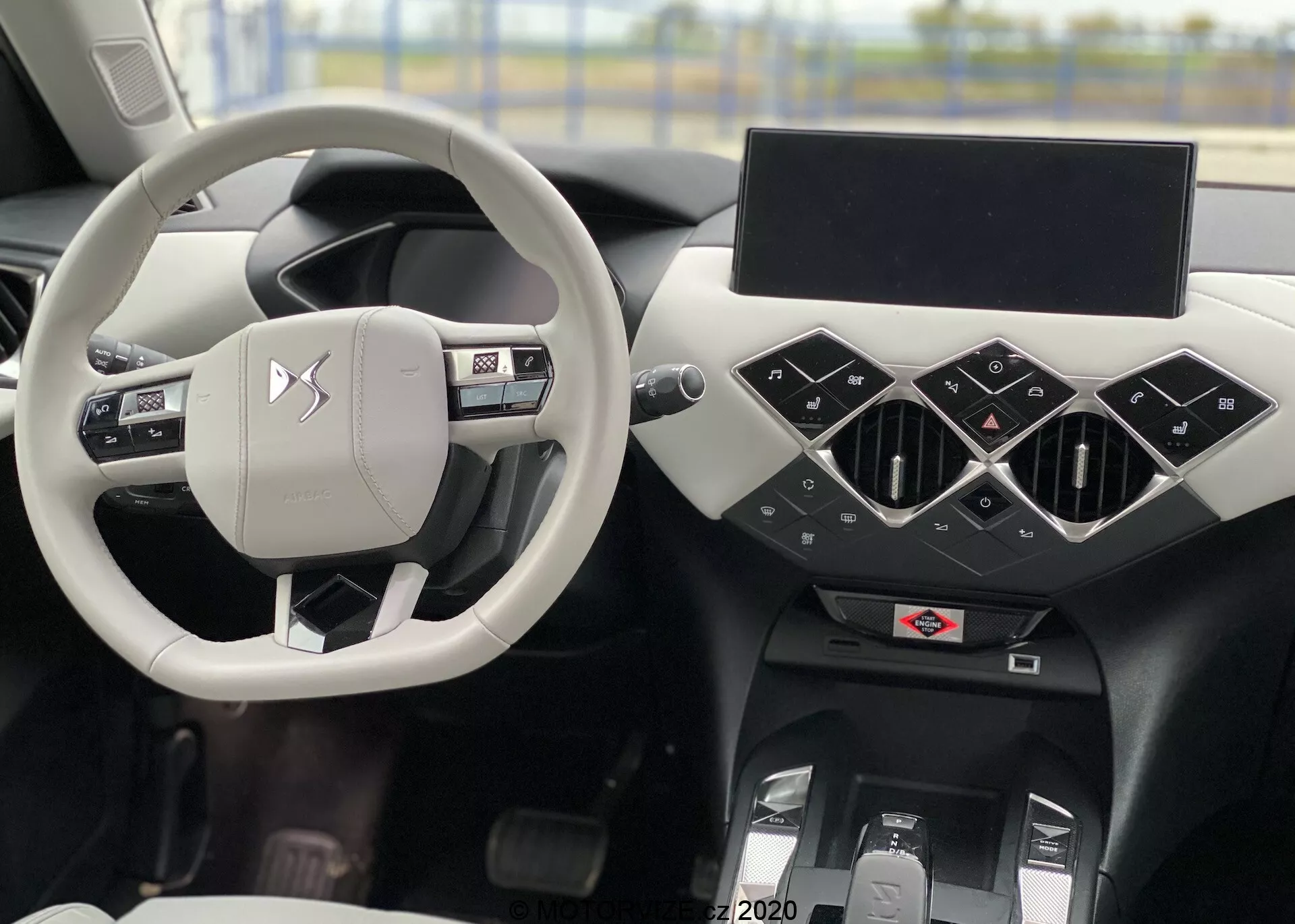 Wnętrze DS Automobiles DS 3 Crossback E-Tense 2019 z centralnie umieszczoną srebrną/jasnoszarą kierownicą z przyciskami sterowania i logo producenta, przez którą widać fotel kierowcy. Czarna deska rozdzielcza ma geometryczne elementy z trójkątnymi centralnymi nawiewami, wyłączony ekran infotainment oraz poniżej możliwe sterowanie klimatyzacją i tunel centralny. Po lewej stronie widać częściowo drzwi kierowcy z przyciskami do obsługi szyb i lusterek.