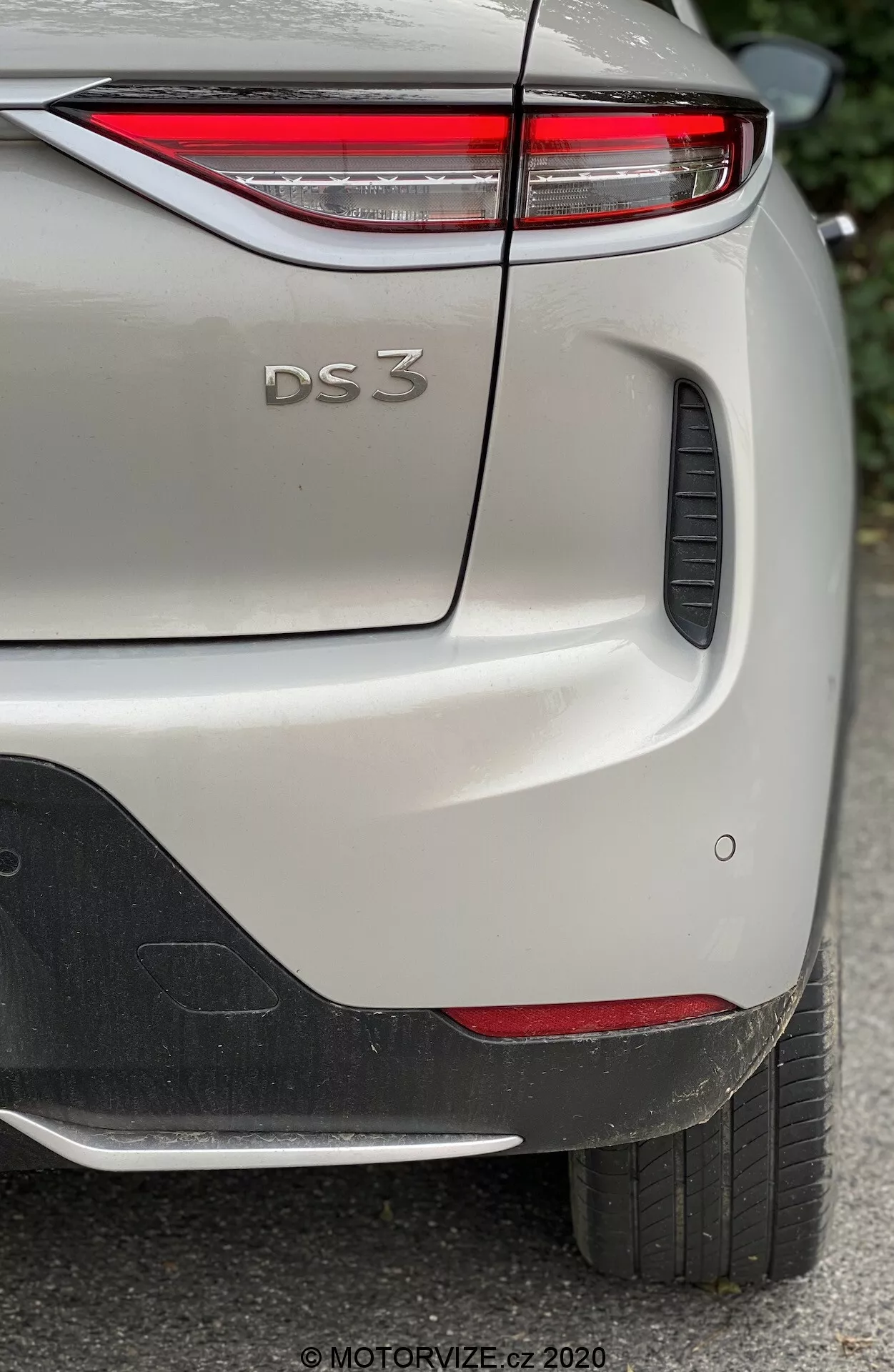 Zbliżenie widoku tylnej bocznej części DS Automobiles DS 3 Crossback E-Tense 2019, koncentrujące się na górnej prawej części tylnego zderzaka, pokrywie bagażnika i części prawego tylnego błotnika. Obraz uchwytuje charakterystyczne detale projektowe modelu, w tym cienkie tylne światła LED, wytłoczone oznaczenie 'DS 3', czujniki parkingowe i czarny plastikowy element na zderzaku. Samochód, pokazany w jasnym odcieniu z metalicznym wykończeniem, jest sfotografowany w naturalnym świetle, uwydatniając drobne detale lakieru i tekstury.