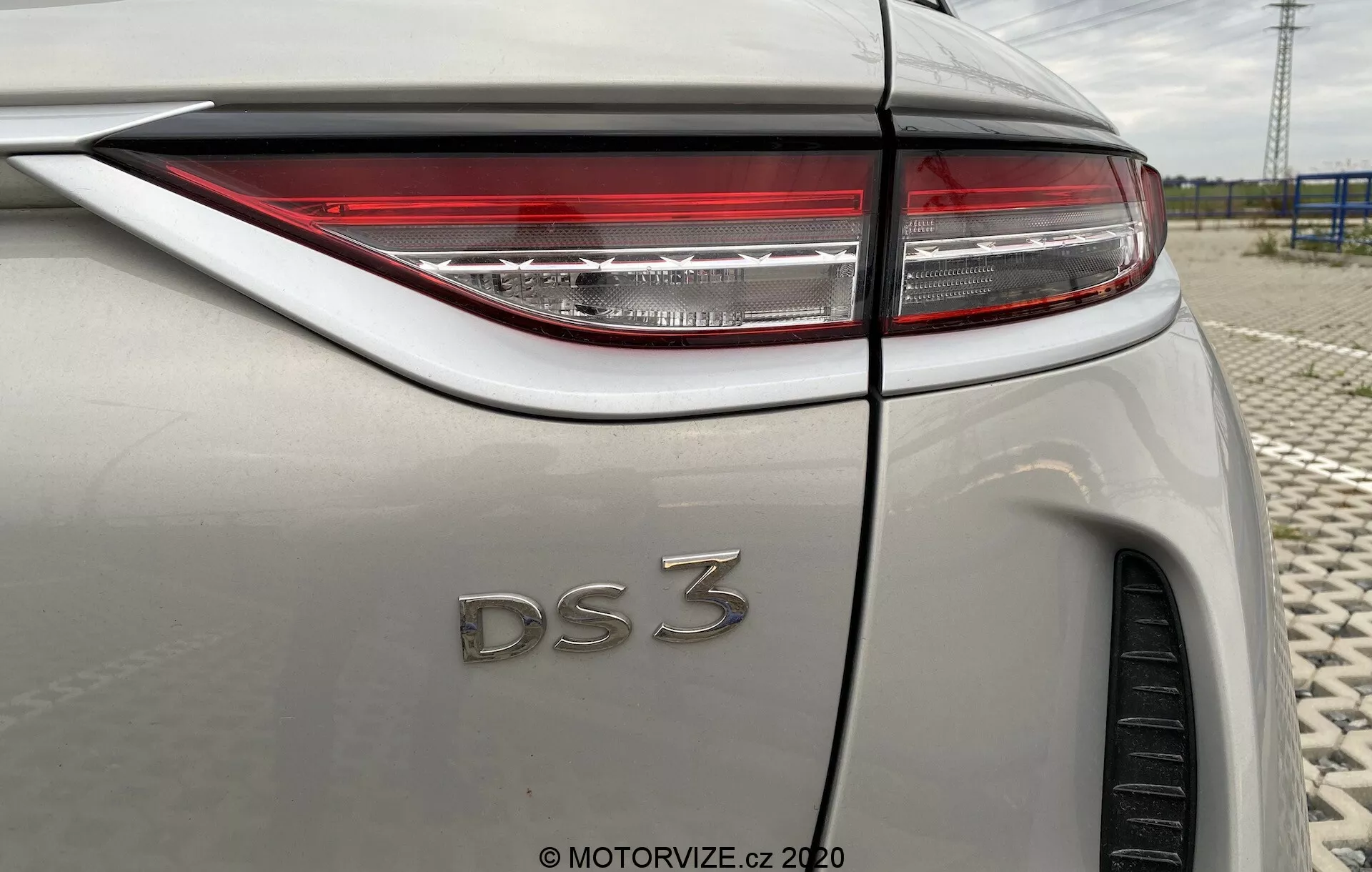 Zbliżenie tylnego trzyczęściowego kąta DS Automobiles DS 3 Crossback E-Tense 2019. Obraz skupia się na prawej stronie pojazdu, podkreślając ostre tylne światła LED, plakietkę 'DS 3' i lekką metaliczną karoserię. Widoczne cechy obejmują linię zagięcia tylnego błotnika, częściową widoczność tylnej opony, antenę typu płetwa rekina i przejście tylnego okna w linię dachu, wszystko pod odbiciem pochmurnego nieba.