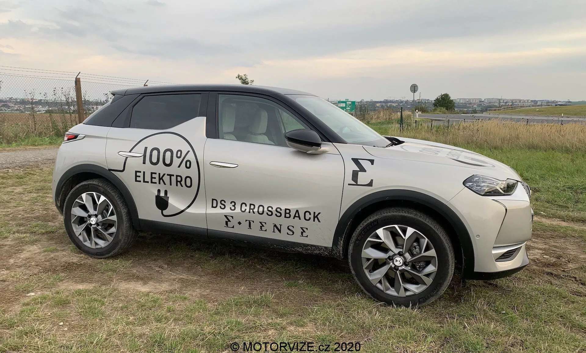 Widok z boku nowoczesnego SUV-a, DS 3 Crossback E-Tense z 2019 roku, pojazdu elektrycznego z napisami '100% ELEKTRO' na boku. Pojazd ustawiony na trawiastym podłożu, z kawałkiem utwardzonej drogi w tle, co pozwala na wyraźne zobaczenie przedniego grilla, świateł LED, przedniego koła i profilu pod lekkim kątem. Widoczne cechy to wieloszprychowe felgi aluminiowe, lusterka boczne z wbudowanymi kierunkowskazami, wpuszczane klamki drzwi oraz symbol portu ładowania elektrycznego na przednim błotniku, sugerujące nacisk na zaawansowaną technologię, styl i ekologiczność, wszystko to w naturalnym, pochmurnym oświetleniu.