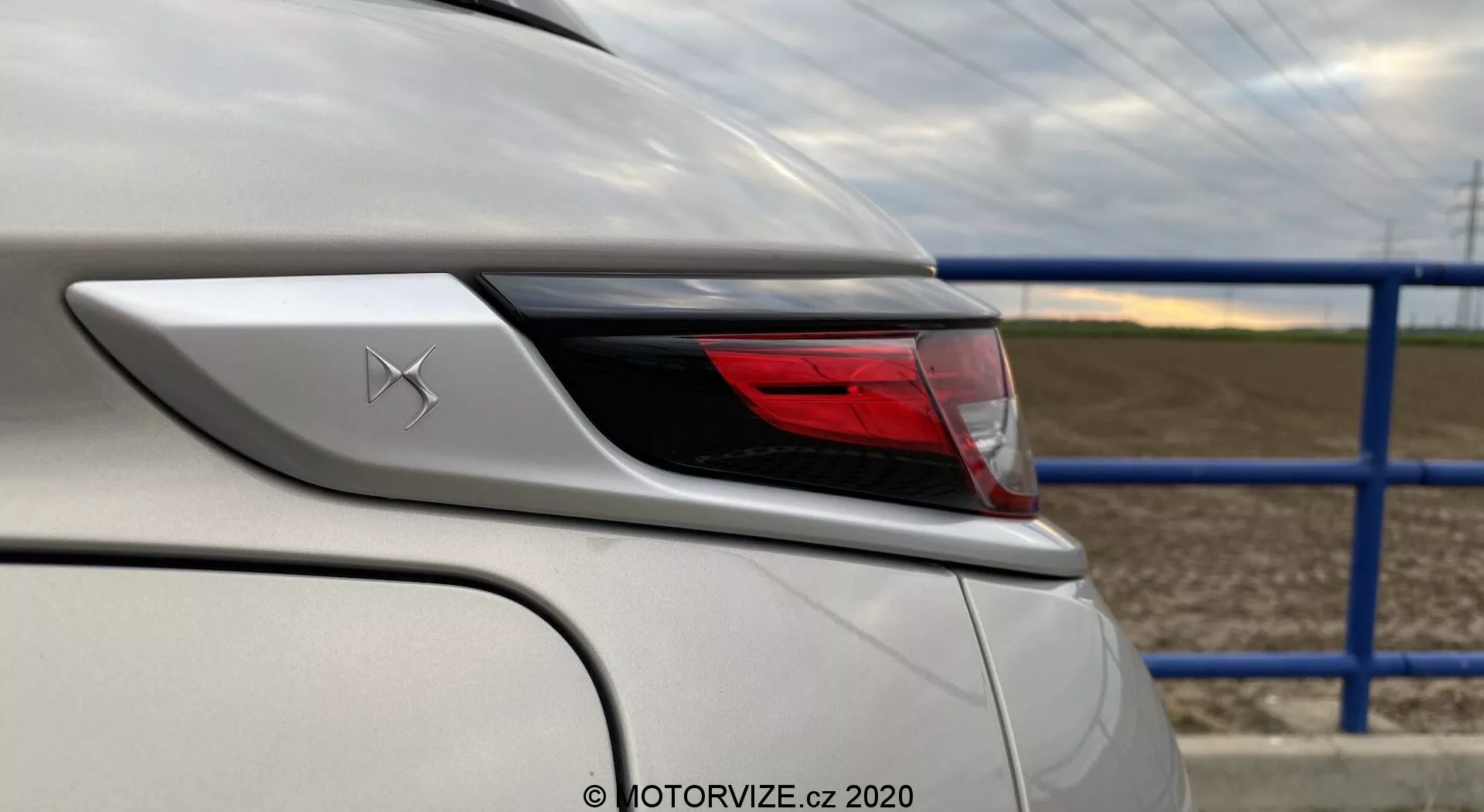 Zbliżenie tylnego rogu białego pojazdu DS-3 2019 (DS-3 Crossback E-Tense), skupiające się na elementach projektu i cechach wokół obszaru tylnego światła. Ujęcie z lewego tylnego narożnika podkreśla krzywiznę tylnego światła i tylnego błotnika, ukazując szczegóły karoserii samochodu oraz zaawansowany zestaw świateł LED. Nad tylnym światłem widoczny jest emblemat ze stylizowanym X, biały lakier w doskonałym stanie daje lśniące wykończenie. W tle znajduje się droga z niebieskim metalowym płotem i polami uprawnymi.