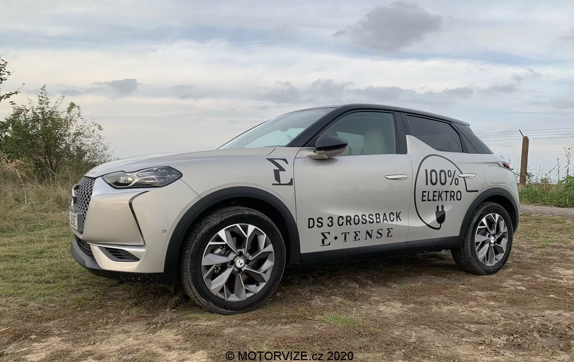Profil boczny elektrycznego kompaktowego SUV-a DS 3 Crossback E-Tense 2019 w jasnym, piaszczystym odcieniu z kontrastowym czarnym dachem. Obraz ukazuje przedni grill z chromowaną obwódką, reflektory LED, odznaki '100% ELECTRO' i 'E-TENSE' z boku, wieloramienne felgi aluminiowe oraz wpuszczane klamki drzwi. Pojazd jest umieszczony na zewnątrz na trawiastym terenie pod zachmurzonym niebem, sugerującym wczesnoporanne lub późne popołudniowe oświetlenie.