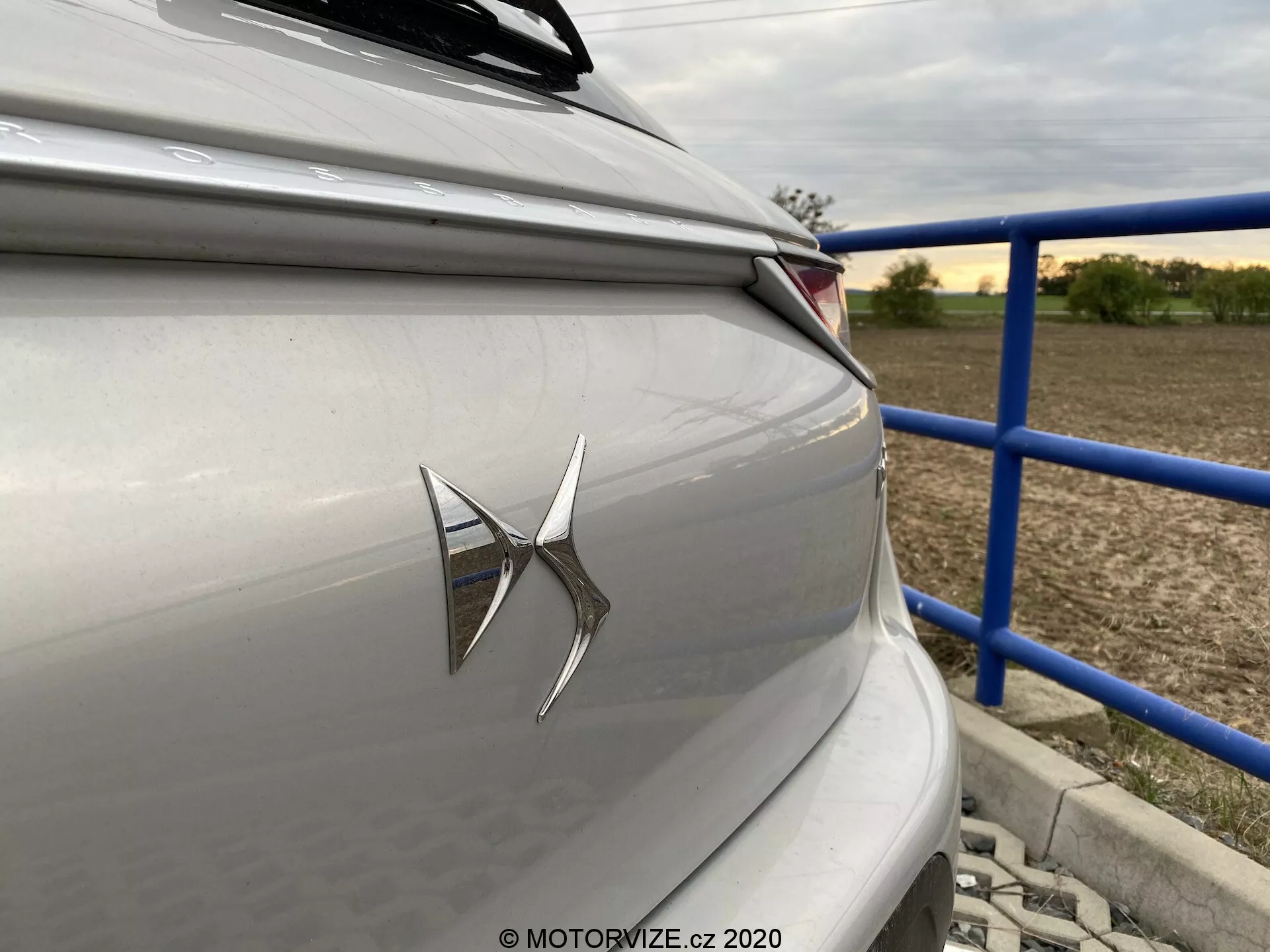 Zbliżenie na tylną część luksusowego sedana lub hatchbacka w jasnym srebrze lub szarym kolorze, z wyraźnym emblematem marki na bagażniku i nowoczesnym klastrem tylnych świateł LED. Zdjęcie, zrobione pod lekkim kątem od strony tylnej, ukazuje ostre poziome linie wzornicze i podmiejskie lub wiejskie tło z niebieskimi poręczami i strukturami. Estetyka wzornictwa samochodu podkreśla współczesną technologię i neutralną, stonowaną kolorystykę.