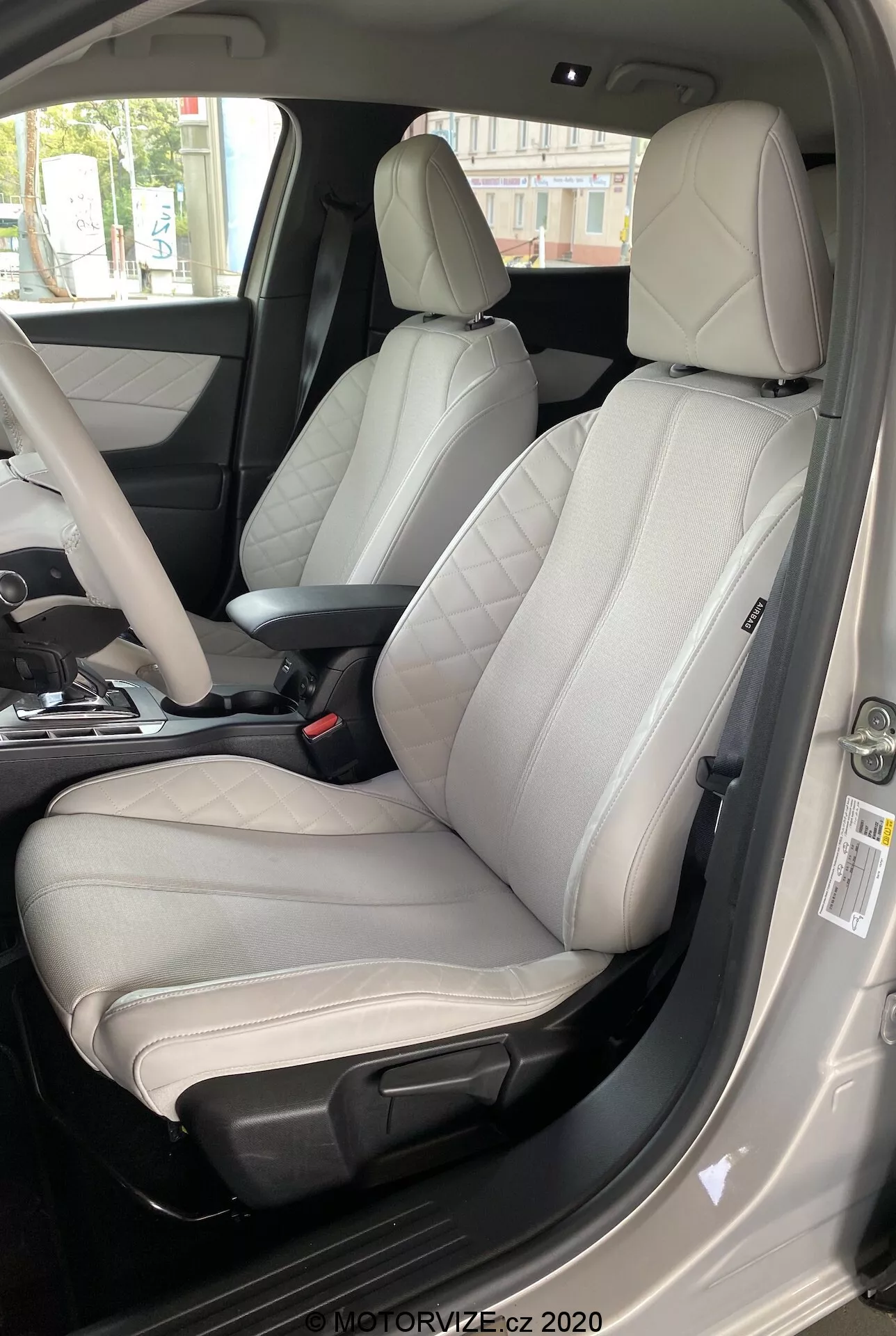 Wnętrze DS Automobiles DS 3 Crossback E-Tense 2019, skupiające się na fotelu kierowcy z perspektywy zewnętrznej po stronie kierowcy. Zdjęcie, wykonane z niskiego na wysoki kąt, pokazuje nowoczesny dwukolorowy skórzany fotel z jasnoszarym środkiem i ciemnoszarymi lub czarnymi krawędziami. Wyraźnie zaprojektowany dla komfortu i sportowej estetyki, fotel ma wyraźne boczne podparcia, widoczne szwy i elektroniczne elementy regulacyjne. Otwórz drzwi kierowcy, widoczny pas bezpieczeństwa, częściowo widoczny środkowy panel oraz panel drzwi z różnymi przyciskami sterującymi wskazują na materiały premium i liczne zaawansowane funkcje. Oświetlenie dzienne zwiększa klarowność luksusowych szczegółów wnętrza.