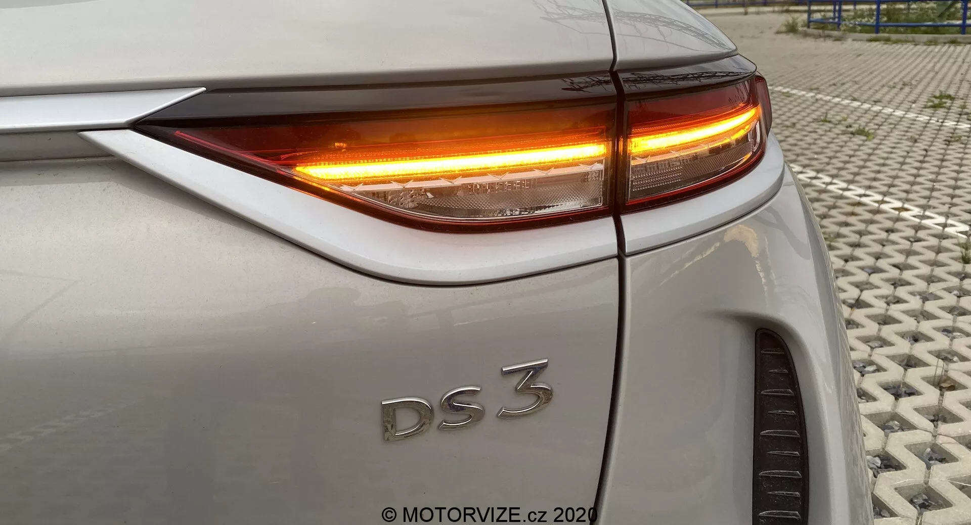 Zbliżenie tylnej części DS 3 Crossback E-Tense (model z 2019 roku), ukazujące prawą tylną ćwiartkę pod delikatnie podwyższonym kątem. Obraz podkreśla tylną lampę LED oraz kierunkowskaz z jasno pomarańczowym oświetleniem. Widoczne elementy obejmują górną krawędź bagażnika, zakrzywienie tylnego błotnika, precyzyjne wyrównanie paneli nadwozia oraz część prawego tylnego koła. Samochód ma srebrny lub jasnoszary lakier. Zdjęcie zrobiono na teksturowanej powierzchni.
