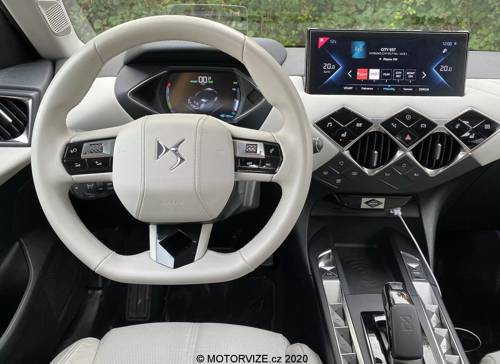Widok wnętrza nowoczesnego samochodu z perspektywy kierowcy, podkreślający wyrafinowane elementy ds-automobiles ds-3 2019 (ds-3-crossback-e-tense). Centralnym punktem jest multifunkcyjna kierownica w jasnym kolorze z przyciskami sterującymi i łopatkami, umieszczona na tle cyfrowego zestawu wskaźników wyświetlającego informacje o pojeździe z jasną, czytelną grafiką. Nad konsolą środkową znajduje się szerokoekranowy, dotykowy wyświetlacz infotainment, otoczony ukośnymi otworami wentylacyjnymi. Centralny tunel mieści automatyczny selektor biegów, sterowanie trybami jazdy oraz dwa uchwyty na napoje. Luksusowe wnętrze o wysokiej jakości charakteryzuje się mieszanką jasnych i ciemnych tonów, symbolizując komfort, wygodę i zaawansowaną technologię motoryzacyjną.