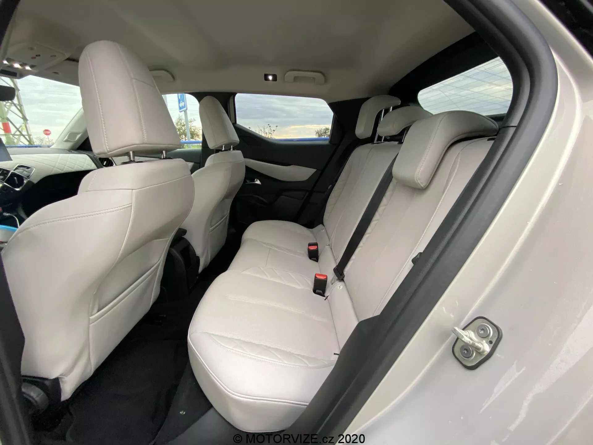 Wnętrze DS Automobiles DS 3 Crossback E-Tense 2019 z nachylonego kąta, koncentrującego się na tylnej części pasażerskiej. Jasne skórzane fotele, widoczne zagłówki, pasy bezpieczeństwa i składany podłokietnik na tylnej kanapie wskazują na luksusowe i zadbane wnętrze. Ciemne dywany kontrastują z jasną tapicerką, a na drzwiach wyraźnie widać sterowanie elektrycznymi szybami. Kąt kamery lekko w dół od tylnego prawego drzwi podkreśla miejsce na siedzenia i przestrzeń na nogi.