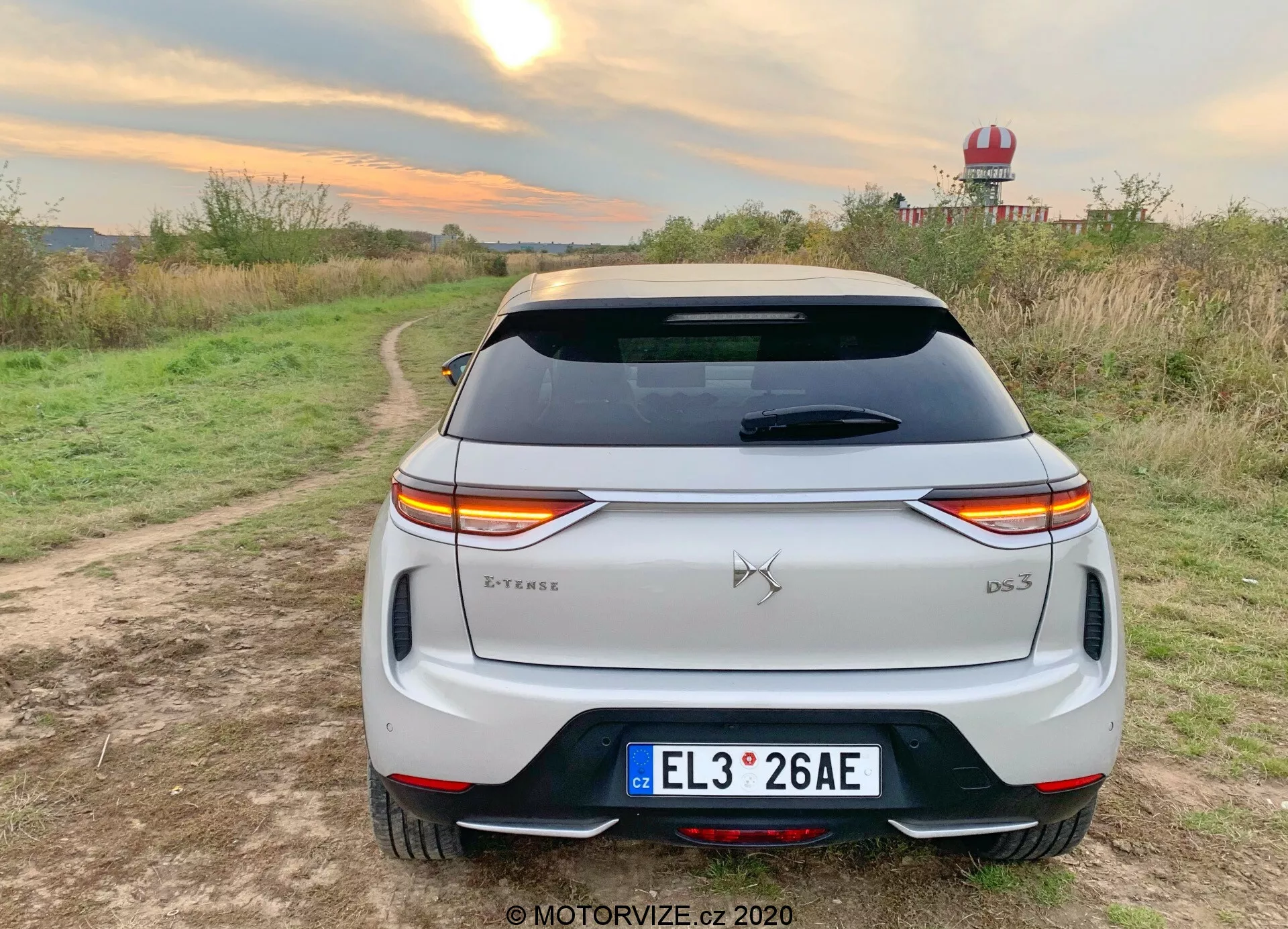 Tylny widok białego crossovera SUV 2019 DS Automobiles DS 3 Crossback E-Tense zaparkowanego na polnej drodze z roślinnością po obu stronach w wiejskim otoczeniu. Charakterystyczne poziome LEDowe światła tylne rozciągają się na całej szerokości tylnej części pojazdu, przerywane przez emblemę marki. Odznaczenie modelu 'DS 3' znajduje się w dolnym prawym rogu klapy bagażnika, a odznaczenie 'E-Tense' w dolnym lewym rogu, wskazujące na napęd elektryczny lub hybrydowy. Funkcje obejmują tablicę rejestracyjną w stylu europejskim, tylną szybę z liniami odmrażania, spojler dachowy, tylny zderzak z wbudowanymi odblaskami i czarnym wykończeniem oraz podwójne końcówki wydechu, co sugeruje sportowy design. Otoczenie sugeruje jazdę próbną lub sesję zdjęciową w stylu życia podkreślającą użytkowanie na świeżym powietrzu, z ciepłymi warunkami oświetleniowymi podkreślającymi białą lakiernię pojazdu.