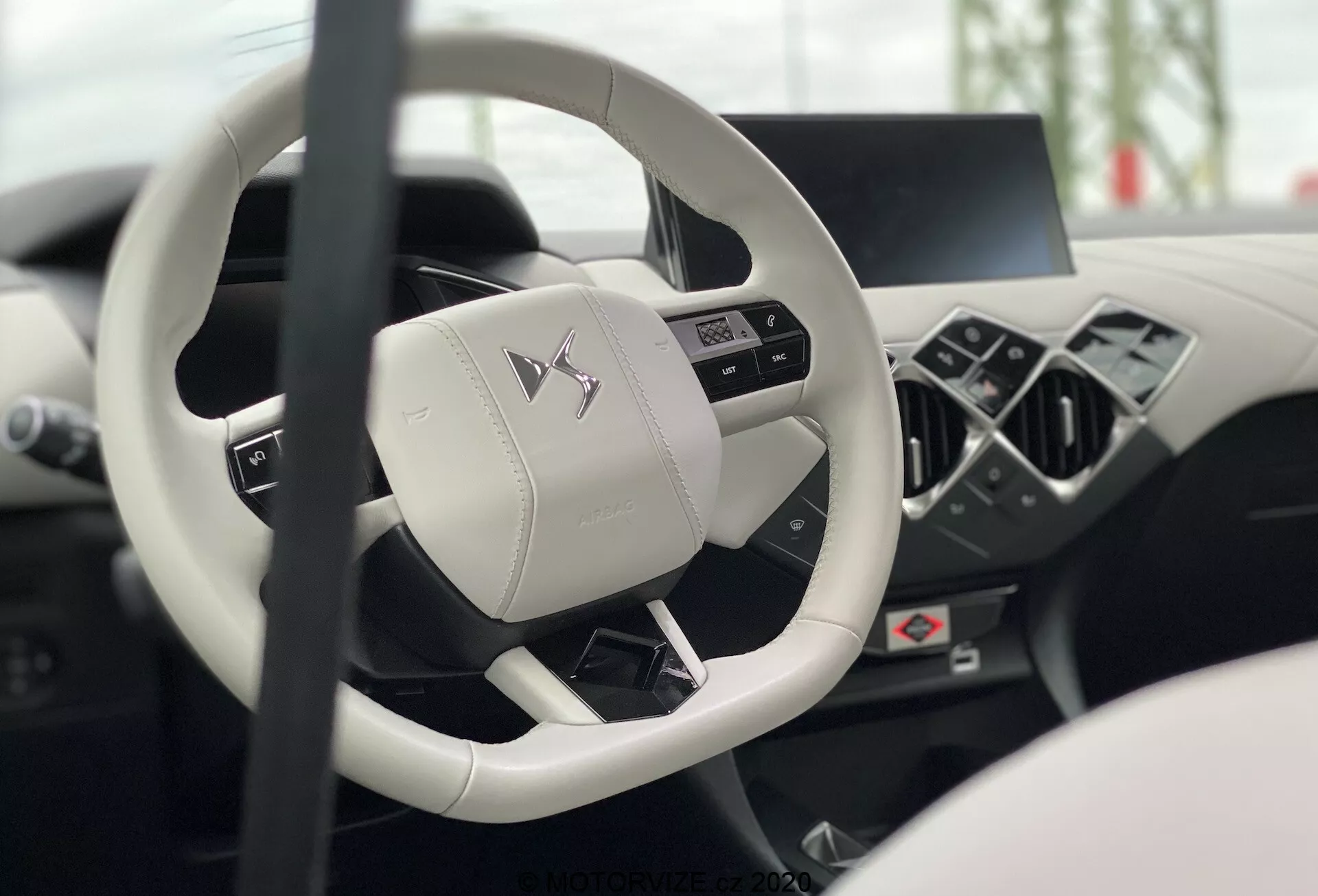 Wnętrze DS Automobiles DS-3 2019 (DS-3-Crossback-E-Tense) z perspektywy siedzenia kierowcy, skupiające się na nowoczesnej trzyramiennej kierownicy z płaskim spodem i wielofunkcyjnymi przyciskami oraz rozmytym cyfrowym kokpicie i konsoli centralnej, ukazujące luksus i zaawansowaną technologię