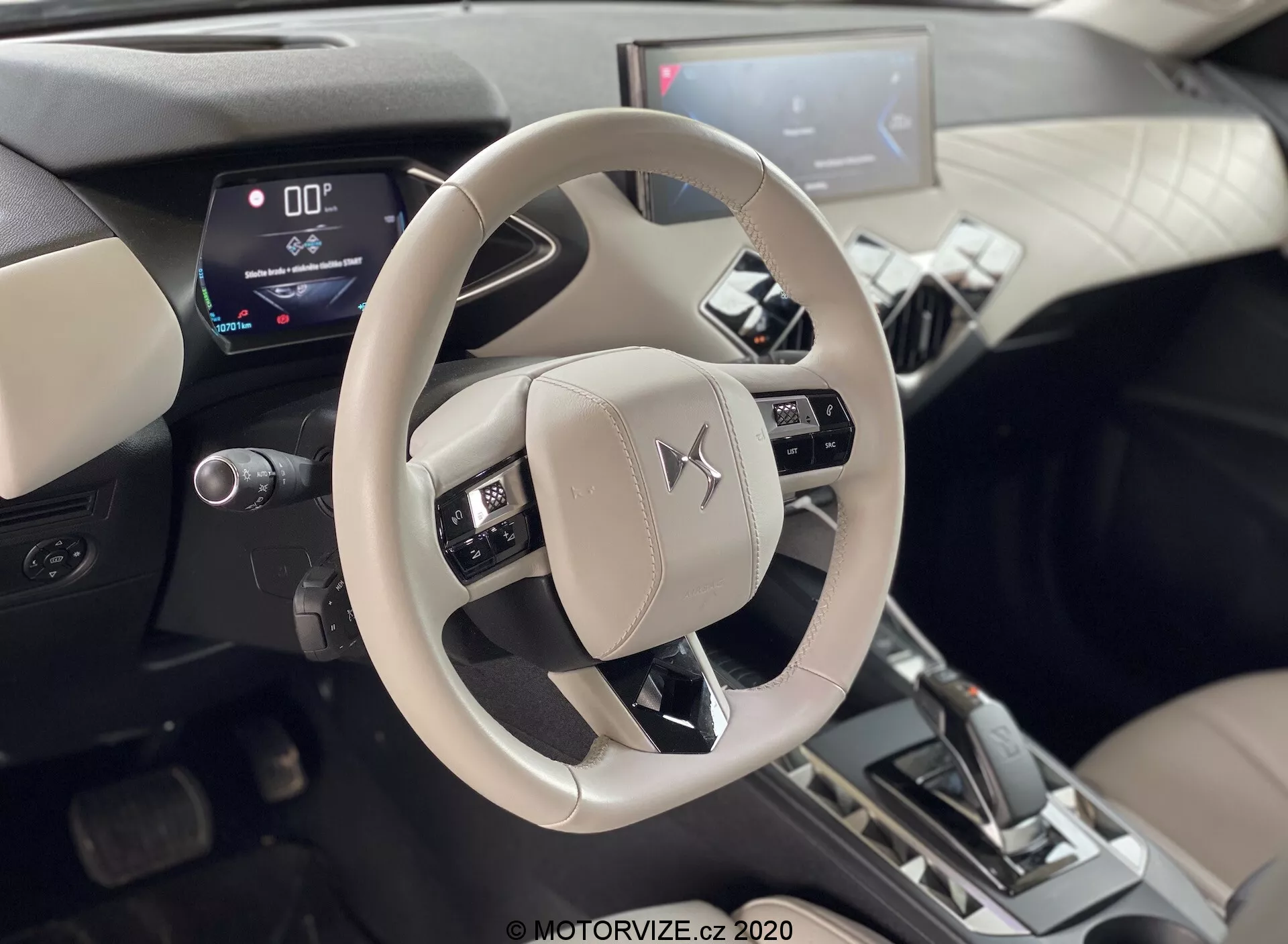 Zdjęcie wnętrza DS-Automobiles DS-3 2019 (DS-3 Crossback E-Tense) skoncentrowane na jasnym kierownicą z emblematem Mitsubishi i konsolą centralną. Zdjęcie z perspektywy osób siedzących z tyłu, ukazujące zamontowane sterowanie dźwiękiem, głosem i systemami kontroli tempomatu. Cyfrowy zestaw wskaźników pokazujący prędkość 0 oraz dźwignia zmiany biegów w pozycji 'P', co sugeruje że pojazd jest na postoju. Widoczny centralny system infotainment z ekranem dotykowym w dwutonowym wnętrzu z metalicznymi akcentami, sugerującymi nowoczesne cyfrowe wyświetlacze i zaawansowane funkcje motoryzacyjne.
