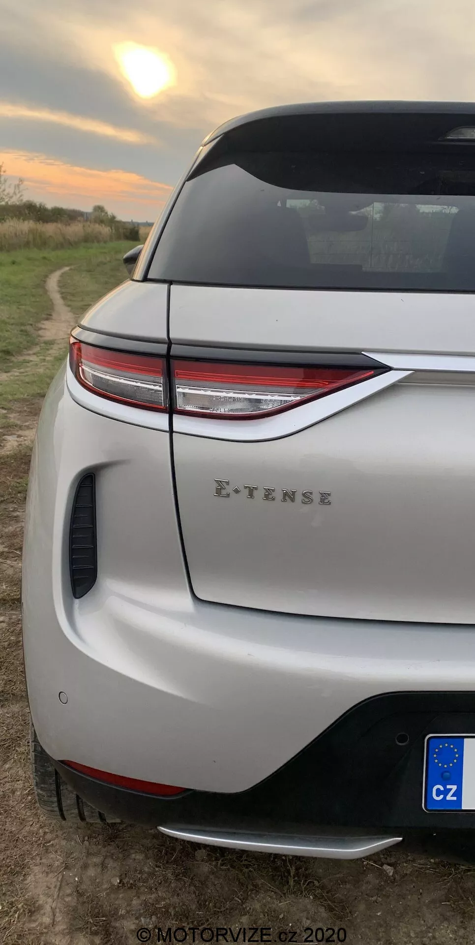 Pionowy zbliżenie tylnego rogu DS-3 2019 (DS-3 Crossback E-TENSE). Skupia się na nowoczesnych tylnych światłach LED, sportowym tylnym zderzaku z wbudowanym dyfuzorem i czujnikiem parkowania, pokrywie bagażnika z aerodynamicznym spojlerem oraz częściowym widoku tylnego koła i błotnika. Ustawione na tle wiejskiego otoczenia z otwartym polem podczas wschodu lub zachodu słońca, wskazując na nowoczesny design pojazdu i jego możliwości elektryczne.