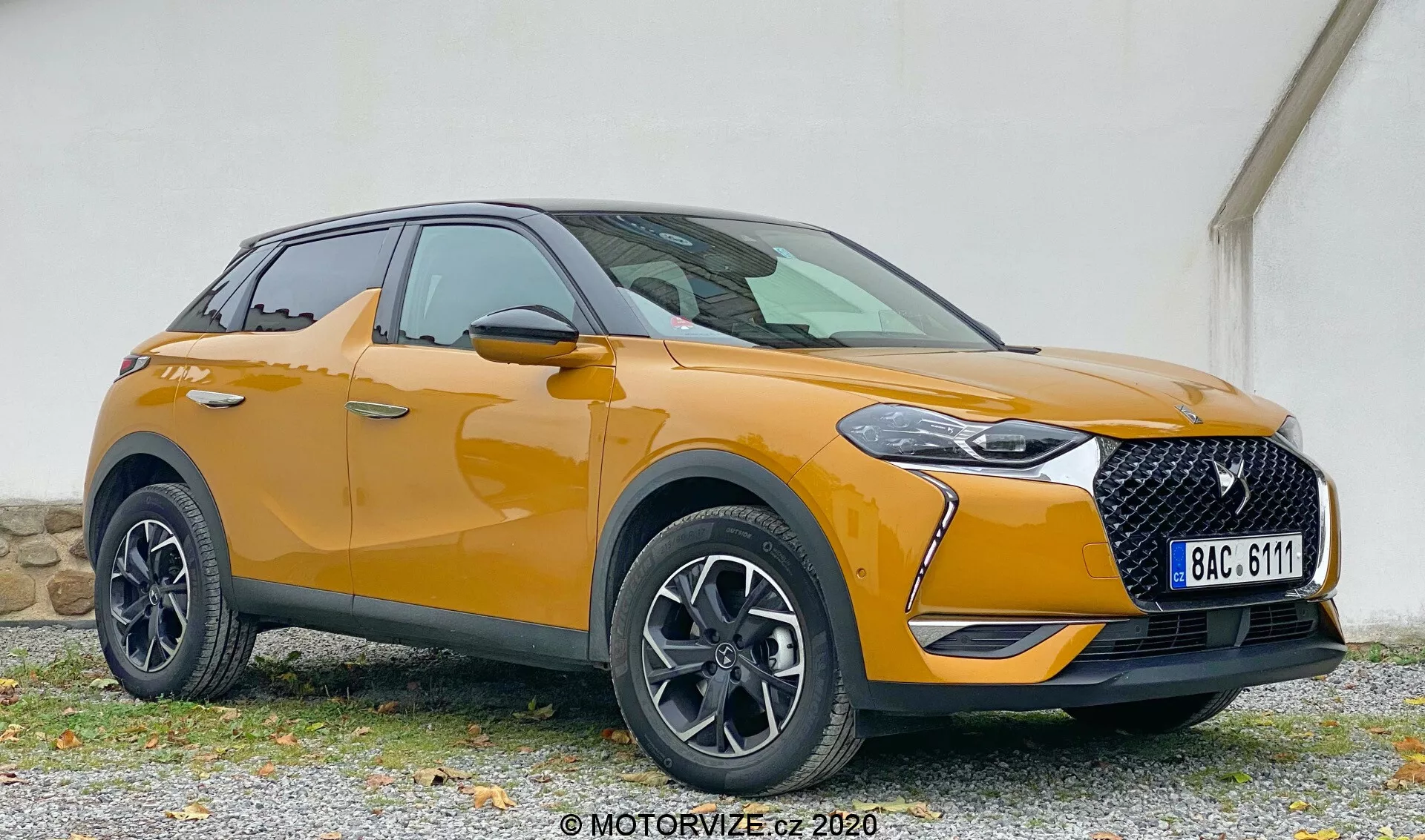 Nowoczesne SUV w widoku przednim pod kątem trzech czwartych pokazujące DS Automobiles DS 3 2019 (DS 3 Crossback) z detalami lewego przedniego rogu, w tym reflektor, kratka, maska, przedni zderzak, koło, przedni błotnik i drzwi kierowcy, ustawione na żwirowej powierzchni na tle o ścianie o stonowanym kolorze.