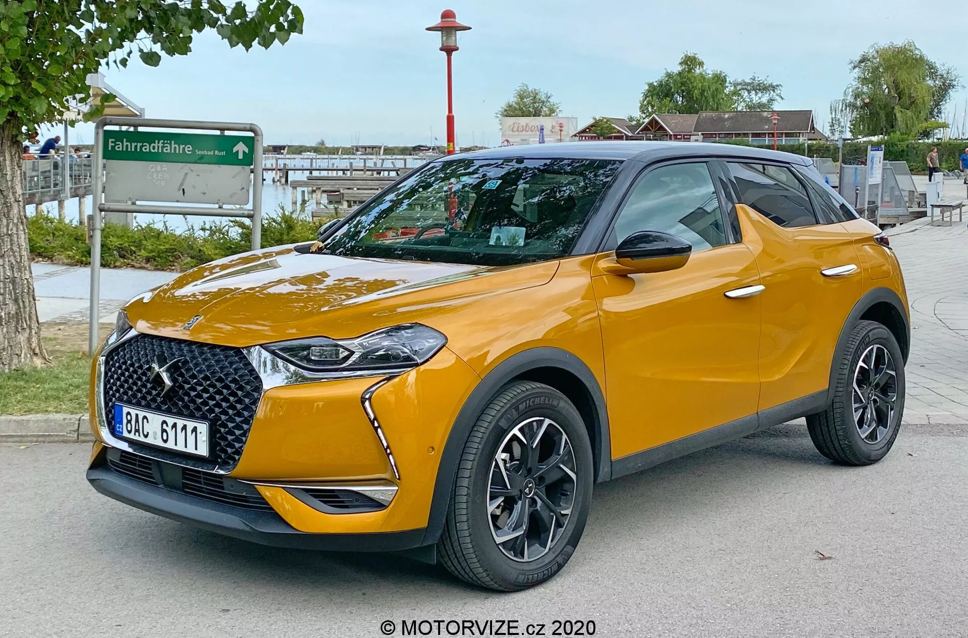 Obraz ukazujący nowoczesne SUV DS Automobiles DS 3 Crossback 2019 w widoku z przedniego trzech czwartych, podkreślający elegancką przednią kratkę, zintegrowane reflektory LED, przedni zderzak, maskę i profil strony kierowcy. Zaparkowany na zewnątrz na utwardzonej powierzchni z miejskim tłem zawierającym stację współdzielenia rowerów. Zdjęcie, zrobione z niskiego punktu widzenia na wysokości reflektorów, akcentuje prześwit samochodu. Widoczne cechy obejmują wieloramienne felgi aluminiowe, lusterka z wbudowanymi kierunkowskazami, opony niskoprofilowe, kontrastowy kolor dachu z relingami dachowymi, metaliczne wykończenie lakieru, specyficzne dla wersji odznaczenia i przyciemnione tylne szyby. Tablica rejestracyjna jest celowo rozmazana dla zachowania prywatności.