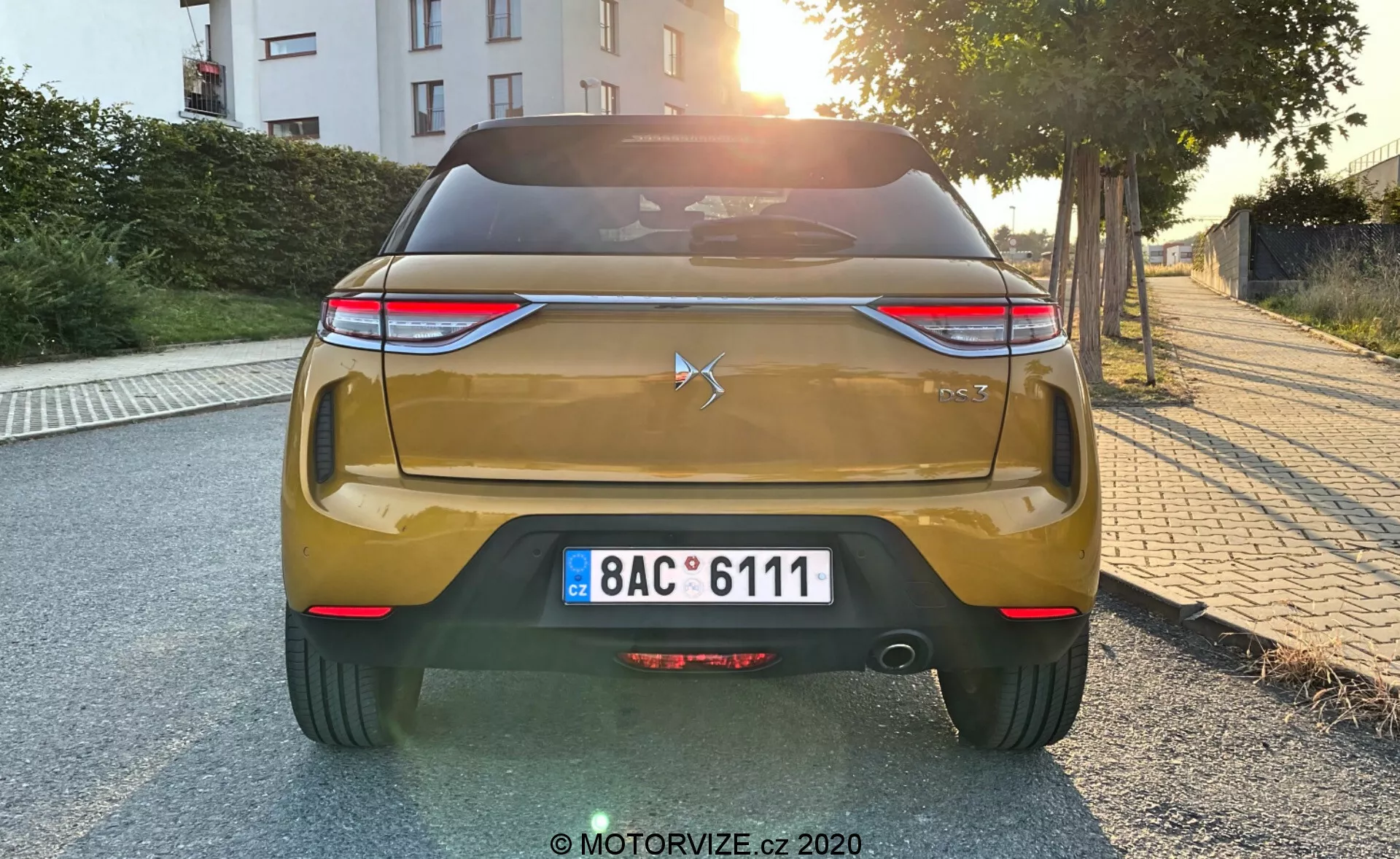 Widok tylnej części złotego samochodu DS Automobiles DS 3 2019 (DS 3 Crossback) o świcie lub zmierzchu. Zdjęcie ukazuje tylne elementy stylizacyjne samochodu, w tym emblemat w kształcie gwiazdy i cyfry '300' na pokrywie bagażnika, poziome tylne światła LED, tablicę rejestracyjną w stylu europejskim, tylni zderzak z odblaskami i czujnikami parkowania, dyfuzor oraz metaliczne wykończenie lakieru.