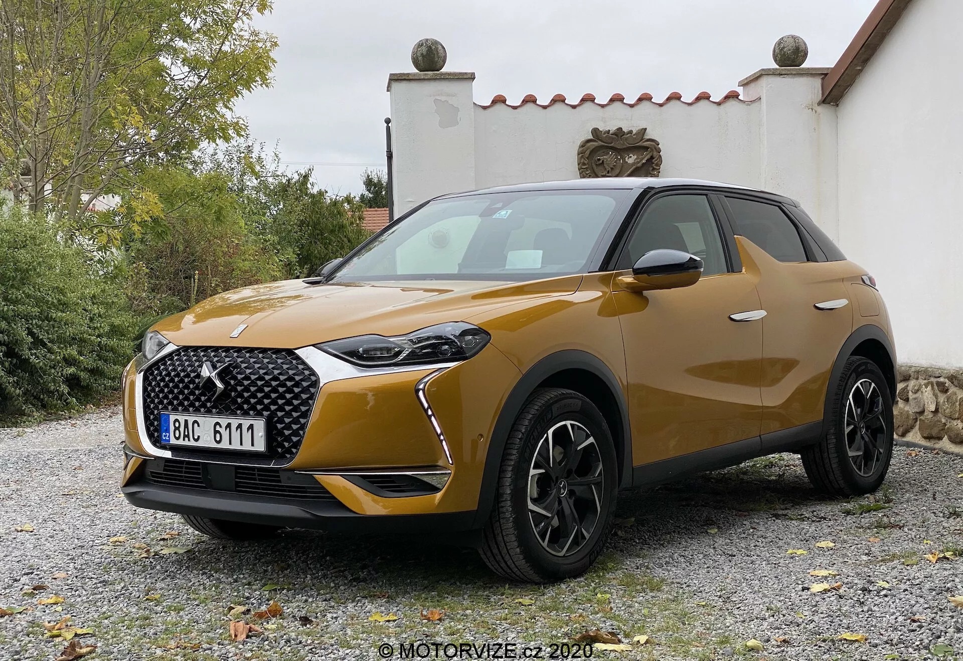 Widok z przodu pod kątem trzech czwartych na DS-3 Crossback z 2019 roku, nowoczesny kompaktowy crossover SUV. Niski kąt podkreśla jego wysokość i mocną sylwetkę pod miękkim i rozproszonym oświetleniem. Kluczowe cechy obejmują charakterystyczne LED-owe światła dzienne otaczające główny zestaw reflektorów, dużą wzorzystą kratkę z wyraźnym znakiem, przyciemnione sekcje reflektorów przeciwmgielnych w zderzaku i zauważalny wlot powietrza pod kratką. Maska ma subtelne kontury, koła są wieloszprychowe czarne aluminiowe, co sugeruje wyższy poziom wykończenia, a boczne lusterka w kolorze nadwozia, prawdopodobnie z zintegrowanymi kierunkowskazami. Płynne linie nadwozia i lekkie wybrzuszenia nad nadkolami nadają wrażenie solidności i sportowego charakteru. Metaliczny lakier wskazuje na poziom premium. Pojazd może posiadać zaawansowane technologie oświetleniowe, czujniki parkowania i być może zintegrowane systemy kamer lub radarów, choć nie są one wyraźnie widoczne. Ustawiony na żwirowej powierzchni z zielenią i budynkiem w tle, widoczna tablica rejestracyjna w stylu europejskim została zmodyfikowana.