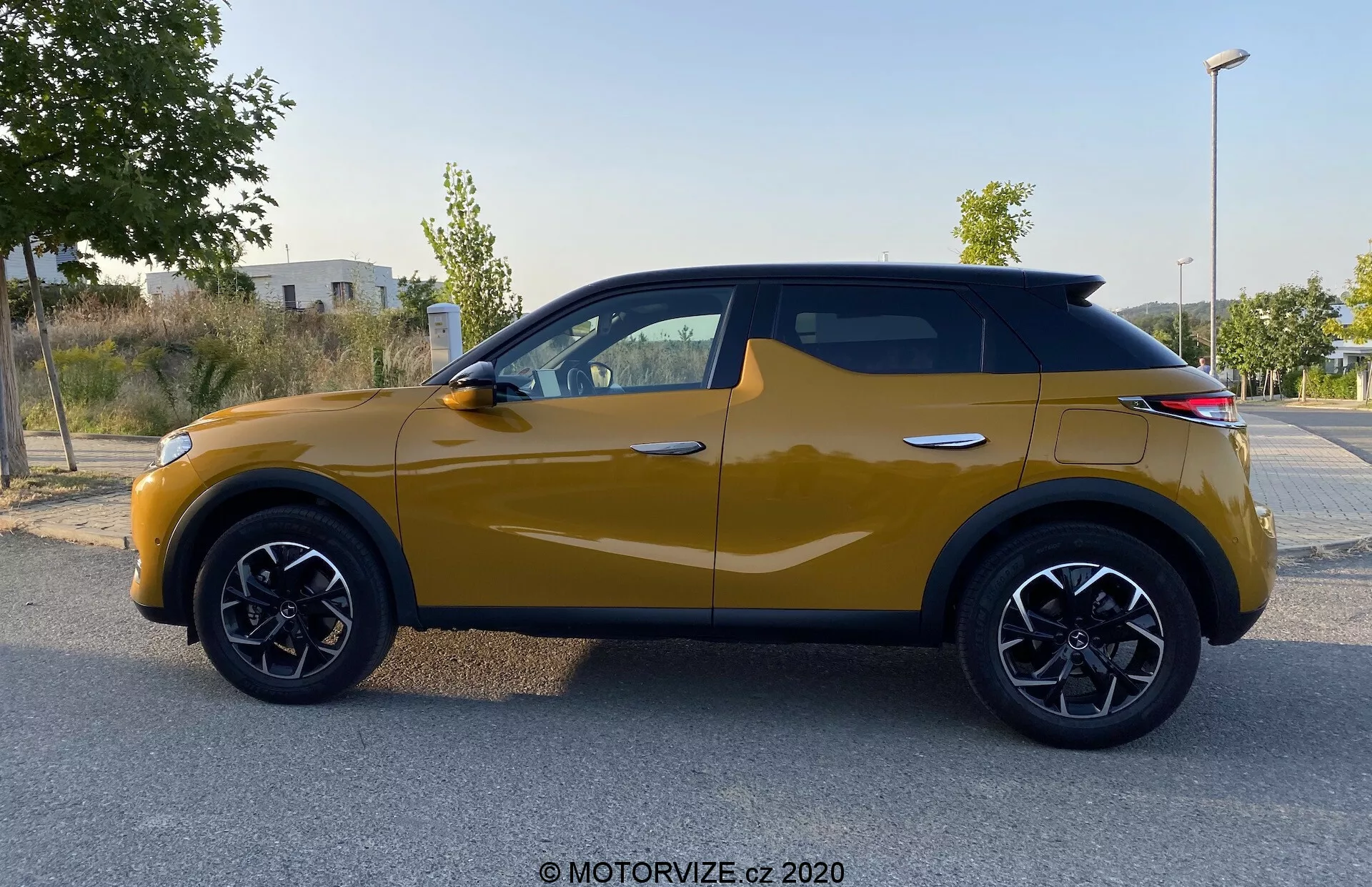 Widok z boku samochodu DS Automobiles DS 3 Crossback 2019 w kolorze musztardowym. Nowoczesny kompaktowy SUV z charakterystycznymi czarnymi nadkolami i czarnymi felgami ze stopu wieloramiennego oraz dynamiczną linią nadwozia od przedniego nadkola po tylne światła. Światła kierunkowskazów wbudowane w lusterka boczne i czarno obramowane okna podkreślają spójny wygląd, a pochylony dach nadaje nowoczesny charakter. Zdjęcie wykonane przy rozproszonym świetle nie zawiera cieni ani odblasków, koncentruje uwagę na elementach designu pojazdu.