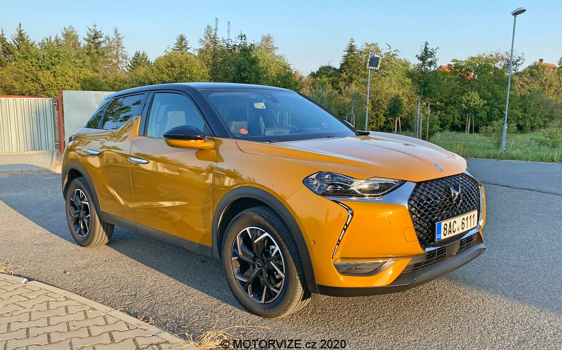 Nowoczesne, kompaktowe SUV DS Automobiles DS-3 Crossback z 2019 roku, pokazane zaparkowane pod kątem z naciskiem na jego profil przedni i boczny. Zdjęcie, uchwycone z niższej perspektywy blisko piast kół, podkreśla jaskrawo pomarańczowy metaliczny korpus pojazdu i czarny dach. Przód zawiera dużą czarną kratkę o wzorze diamentowym, ostre reflektory z akcentami LED i światła przeciwmgielne w czarnych oprawach. Samochód ma czarne lusterka boczne, chromowane listwy okienne i czarne koła ze srebrnymi akcentami. Zwraca uwagę lekko podwyższona postura, sportowe linie nadwozia i fałd na drzwiach opadający ku przodowi.
