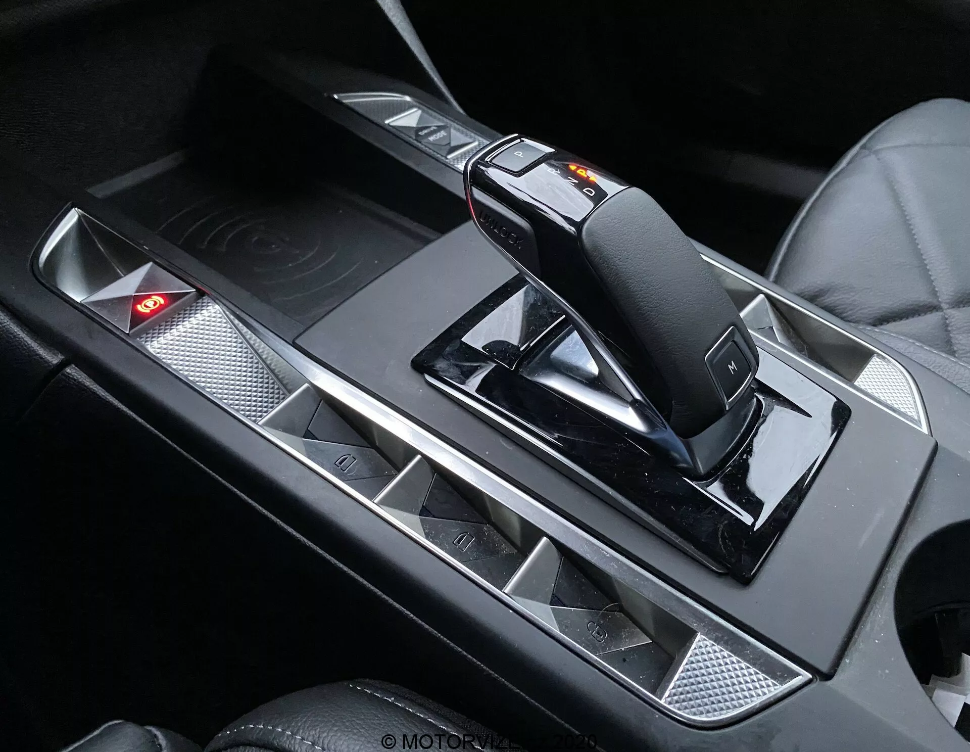 Zbliżenie wnętrza DS Automobiles DS-3 Crossback 2019, skupiające się na konsoli środkowej z nowoczesną, ergonomiczną dźwignią zmiany biegów z przełącznikiem trybu manualnego i mechanizmem zwalniającym. Konsola posiada metaliczne akcenty kontrastujące z czarnymi powierzchniami, uchwyty na kubki z matowym wykończeniem oraz panel sterowania z czerwoną kontrolką ostrzegawczą.