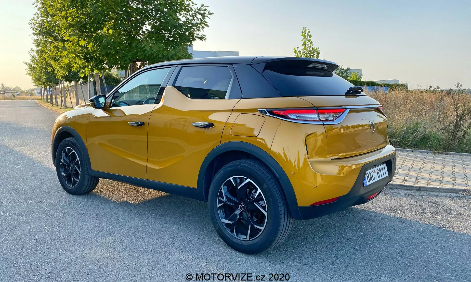 Widok tylny w trzech czwartych nowoczesnego SUV-a crossover w naturalnym świetle dziennym, z żywym metalicznym złotym kolorem i sportowym czarnym dachem, nowoczesnymi tylnymi światłami LED, dużymi czarnymi felgami aluminiowymi, wysokim prześwitem, widocznymi tylnymi czujnikami parkowania i dwukolorowym designem, charakterystycznym dla DS Automobiles DS 3 Crossback z 2019 roku.