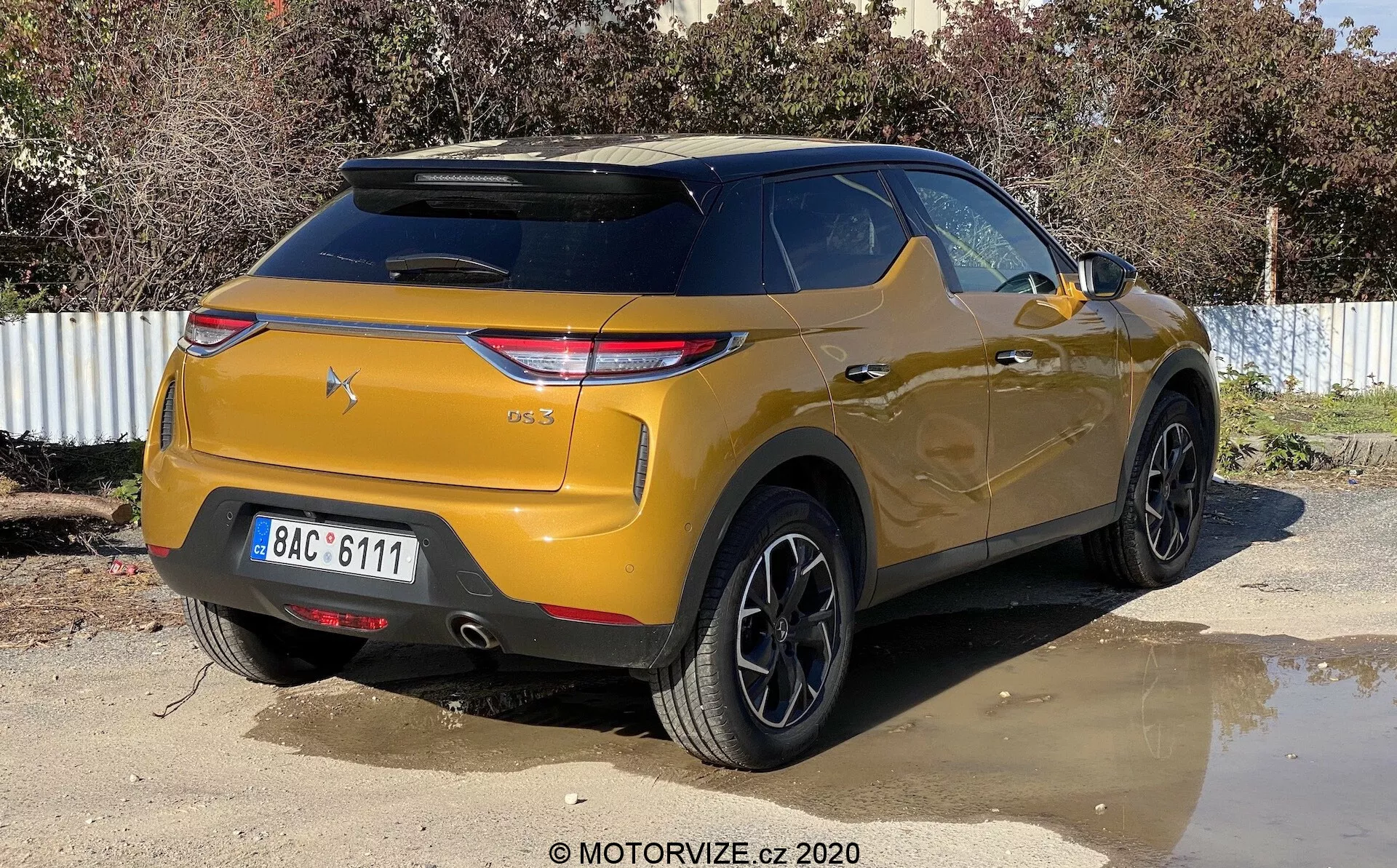 Widok trzej-czwarty z tyłu metalicznego złotego SUV-a DS-3 Crossback zaparkowanego na zewnątrz w słoneczny dzień. Obraz ukazuje pojazd pod lekkim prawym kątem, podkreślając prawą tylną stronę i obszar bagażnika. Kluczowe cechy obejmują nowoczesne, kanciaste tylne światła, pochyloną tylną szybę z wbudowanym spojlerem dachowym, wyraźny zderzak z czujnikami parkowania, czarne wieloszprychowe felgi aluminiowe, wysokie prześwity i przyciemniane tylne szyby.