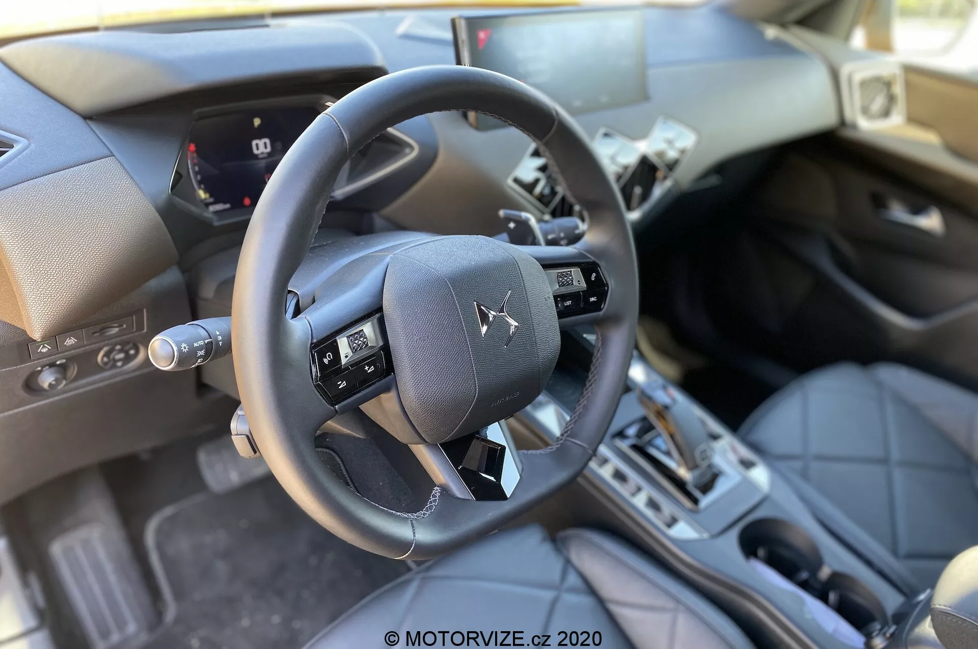 Widok wnętrza DS Automobiles DS 3 Crossback 2019 z perspektywy przedniego siedzenia pasażera, podkreślający trójramienną kierownicę z wielofunkcyjnymi przyciskami, częściowy cyfrowy zestaw wskaźników pokazujący prędkość auta i mierniki, konsolę centralną z dźwignią zmiany biegów automatycznej skrzyni biegów, materiały premium takie jak skórzane powierzchnie i metalowe akcenty, tradycyjny slot zapłonu, pedały oraz dobrze oświetlone naturalnym dziennym światłem.