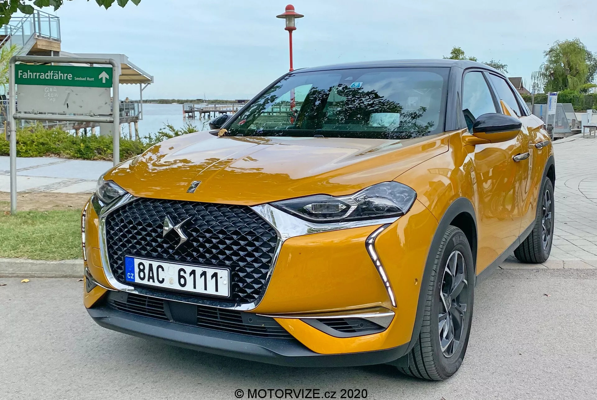 Obraz przedstawia DS Automobiles DS 3 Crossback 2019, nowoczesny kompaktowy crossover SUV, zaparkowany na brukowanej drodze. Uchwycony pod niskim kątem z przodu, obraz ukazuje przedni pas pojazdu i jego prawy bok. Kluczowe cechy obejmują wyraźne logo marki na kratce z wzorem rombu, eleganckie reflektory LED z stylizowanymi światłami do jazdy dziennej, zintegrowane światła przeciwmgielne w przednim zderzaku, lusterka boczne w kolorze nadwozia z kierunkowskazami i ciemne, wieloramienne felgi aluminiowe. Samochód jest pomalowany na intensywny metaliczny pomarańczowy kolor z wyraźnymi liniami nadwozia, które poprawiają jego aerodynamiczny wygląd, oraz oznaczeniami mogącymi wskazywać poziomy wyposażenia. Otoczenie sugeruje obszar publiczny w pobliżu parku, na co wskazuje znak ścieżki rowerowej w tle.