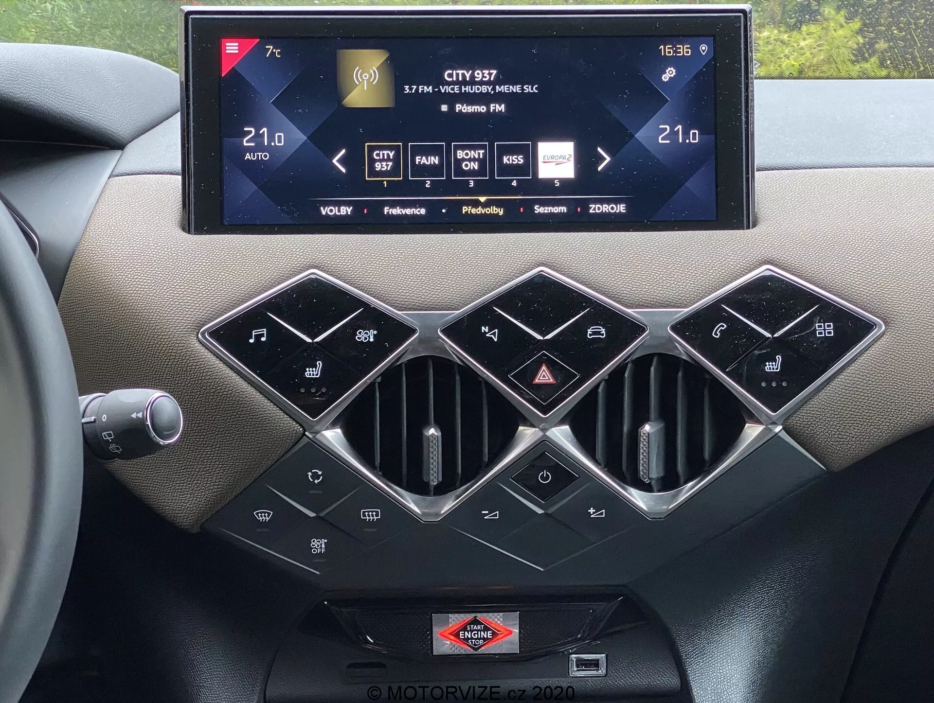 Widok wnętrza konsoli centralnej ds-automobiles ds-3 2019 z dużym ekranem dotykowym systemu infotainment pokazującym sterowanie klimatyzacją, informacje o radiu i ustawienia, z trzema heksagonalnymi otworami wentylacyjnymi z srebrnymi obwódkami poniżej. Poniżej znajduje się rząd czarnych, błyszczących przycisków do sterowania klimatyzacją, w tym przycisk SYNC do sterowania klimatyzacją wielostrefową, funkcje automatycznego sterowania klimatyzacją i plakietka marki umieszczona pod ekranem dotykowym, podkreślająca wysokiej jakości materiały i nowoczesną technologię samochodową