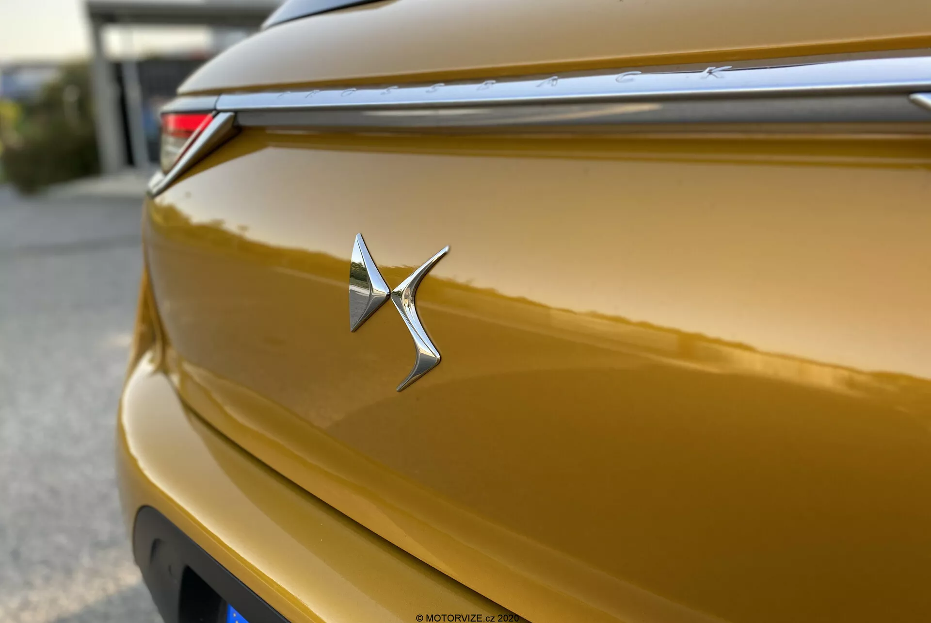 Zbliżenie na metaliczną złotą nasadkę DS Automobiles DS-3 Crossback 2019. Chromowany emblemat jest ostry i centralnie umieszczony, przypominający gwiazdę z wydłużonym prawym ramieniem. Błyszcząca karoseria odbija światło na swoich krzywiznach, pokazując bogaty złoty odcień. Płytka głębia ostrości podkreśla nasadkę, z poziomą linią nad nią, która przyczynia się do aerodynamicznego designu. Częściowo widoczna jest tylna lampa LED w prawym górnym rogu. Pokrywa tylna ma subtelną krzywiznę, a efekt bokeh w tle izoluje nasadkę.