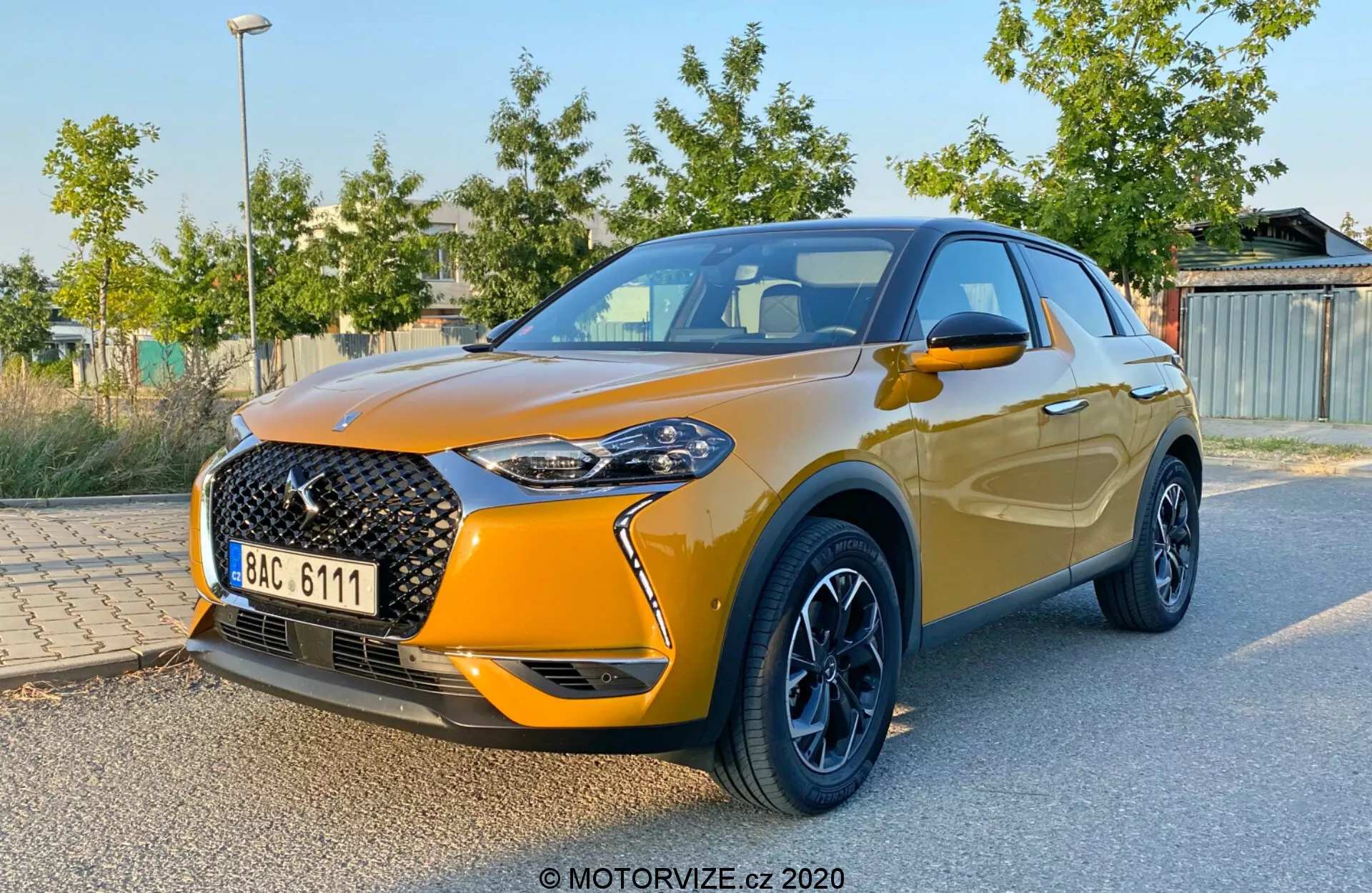 Widok przedni trzy czwarte DS Automobiles DS 3 Crossback 2019, ukazujący charakterystyczną dużą przednią kratkę z wzorem siatki i wyraźnym emblematem, eleganckie reflektory LED, metaliczne pomarańczowe lakierowanie, kontrastowe czarne felgi aluminiowe, zintegrowane lusterka boczne i wznoszącą się linię okien kończącą się aerodynamicznym spojlerem. Pojazd jest zaparkowany z zielonym listowiem i konstrukcjami w rozmytym tle, podkreślając design i sportowe cechy samochodu.