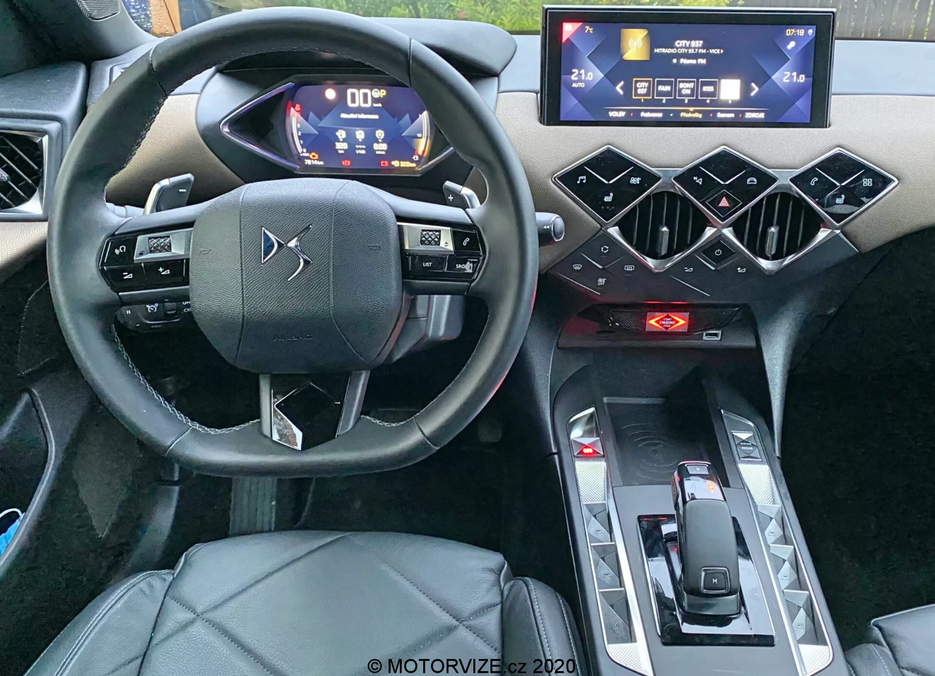 Widok wnętrza DS Automobiles DS-3 Crossback 2019, skupiającego się na kokpicie kierowcy z perspektywy poziomu oczu. Zdjęcie wykonane z tylnej części pasażerskiej ukazuje kierownicę trzyramienną z dotykowymi przyciskami, cyfrowy zestaw wskaźników z niebieskimi i białymi podświetlanymi wskaźnikami, poziomo orientowany system infotainment z ekranem dotykowym, geometryczne, sześciokątne elementy designu na konsoli środkowej, czerwone wskaźniki kontroli klimatu, minimalistyczny selektor biegów oraz fotel kierowcy z tapicerką ze skóry lub skórzanego ożebrowania. Zdjęcie podkreśla nowoczesne, zaawansowane technologicznie funkcje i luksusowy design samochodu.