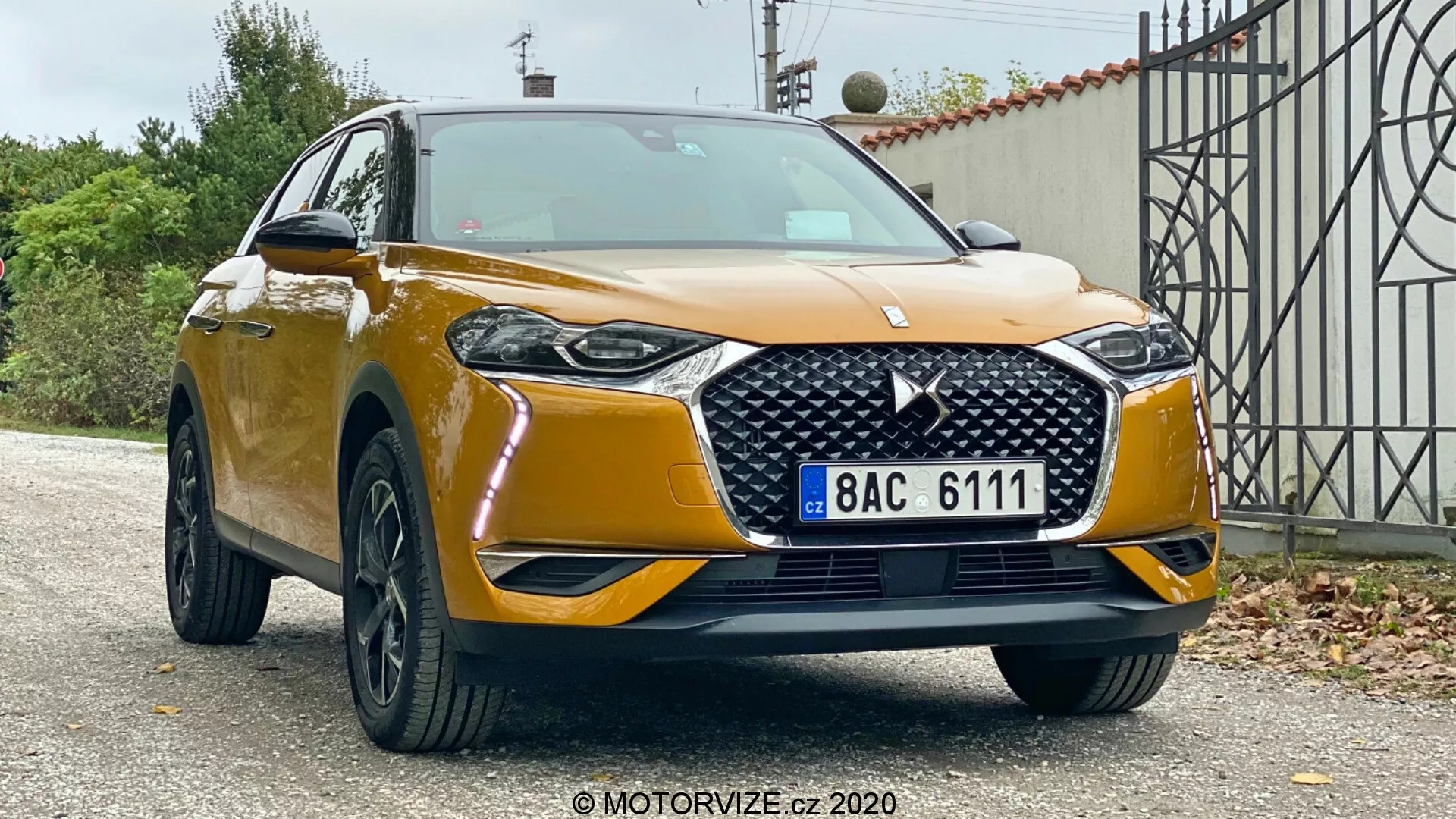 Widok przedni trzy czwarte SUV-a DS Automobiles DS 3 Crossback z 2019 roku, ukazujący jego wysokość i solidną postawę na tle przedmieścia. Widoczne są: metaliczny złoty lakier, wyraźny grill z wzorem diamentu i emblematem, smukłe reflektory LED, światła przeciwmgielne, duży przedni zderzak z szerokim wlotem powietrza, czarne, wieloramienne felgi aluminiowe oraz lusterka boczne z zintegrowanymi kierunkowskazami.